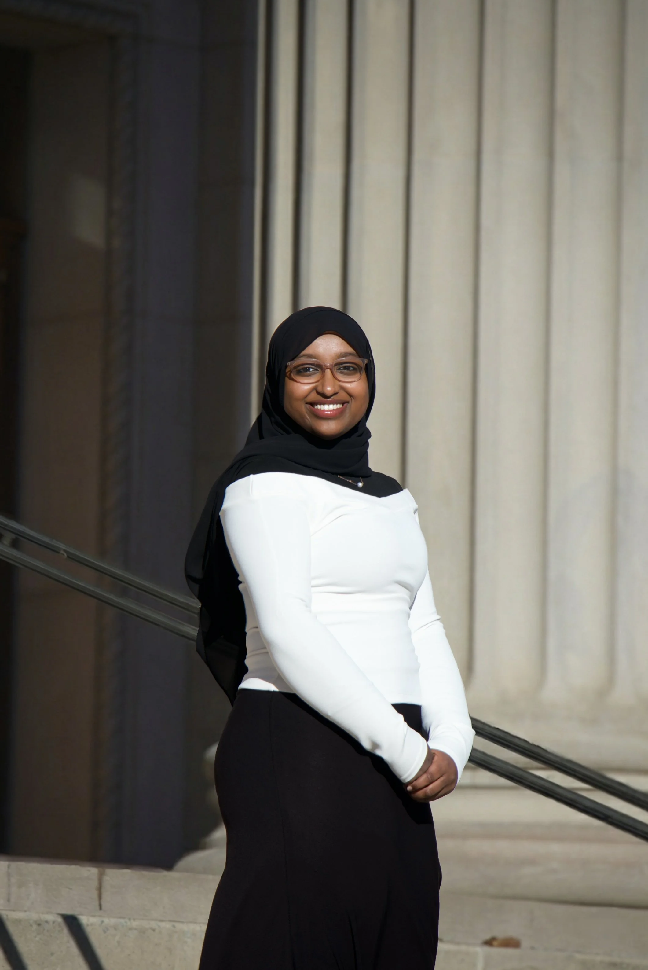 A woman wearing a black hijab, glasses, a white long sleeve top, and a black skirt, smiling and standing outdoors near stairs with a railing.
