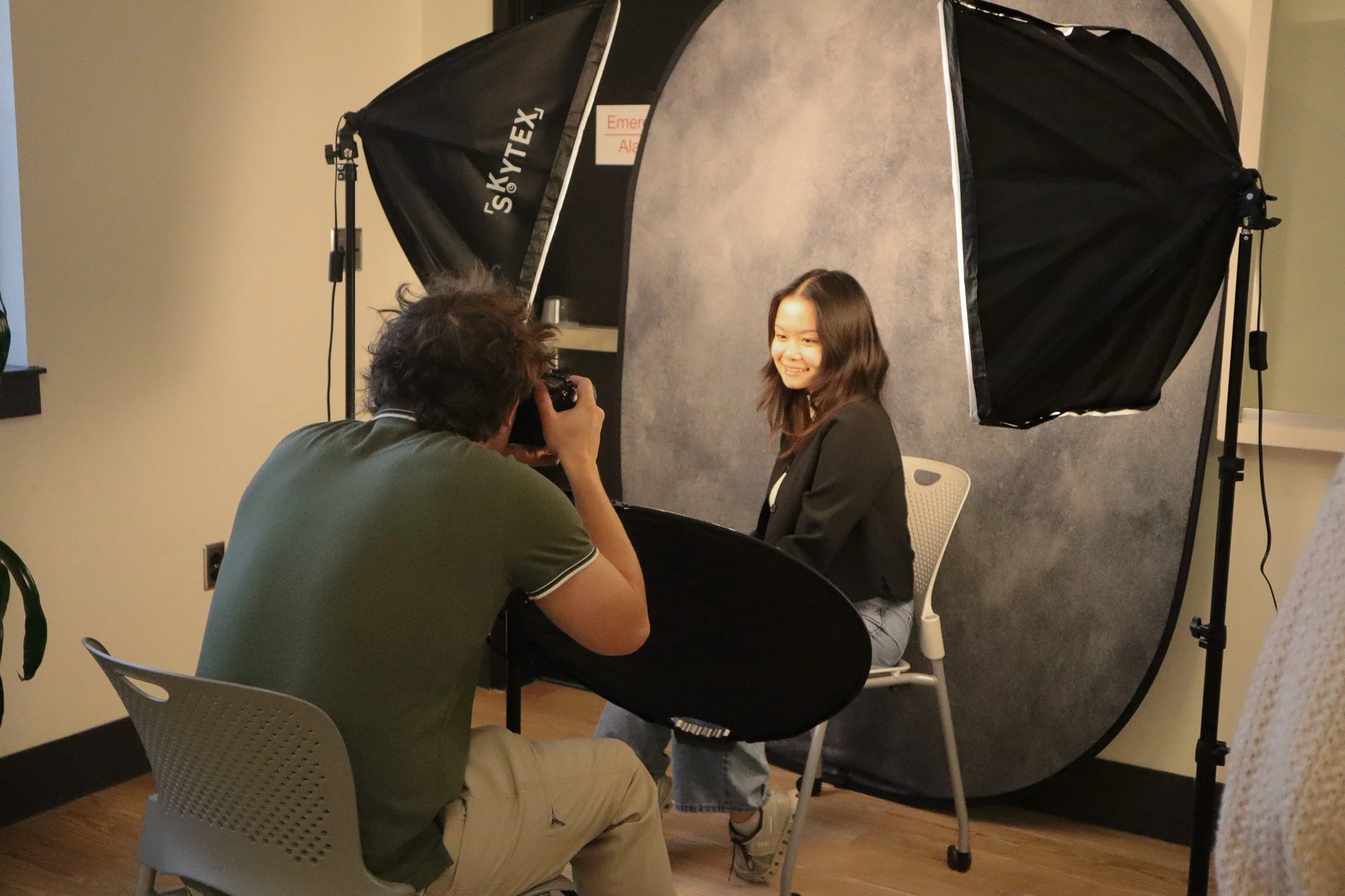 A young woman with long brown hair sits smiling in front of a photography backdrop while a photographer takes her picture during a photo shoot. The setup includes a large black umbrella light and a small stool.