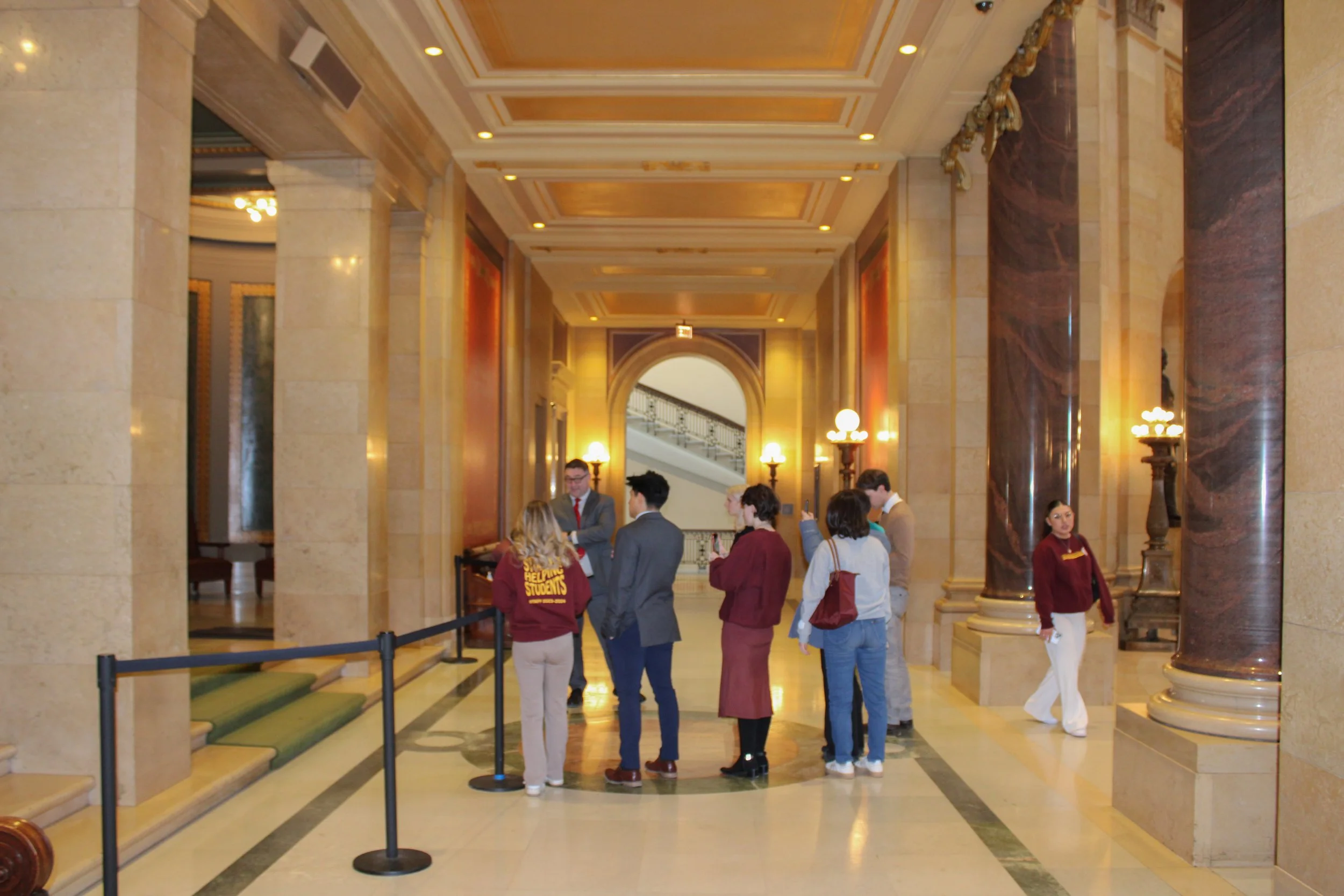 People standing in line and talking in the lobby of a grand, ornate building with marble columns, decorative ceiling, and warm lighting.