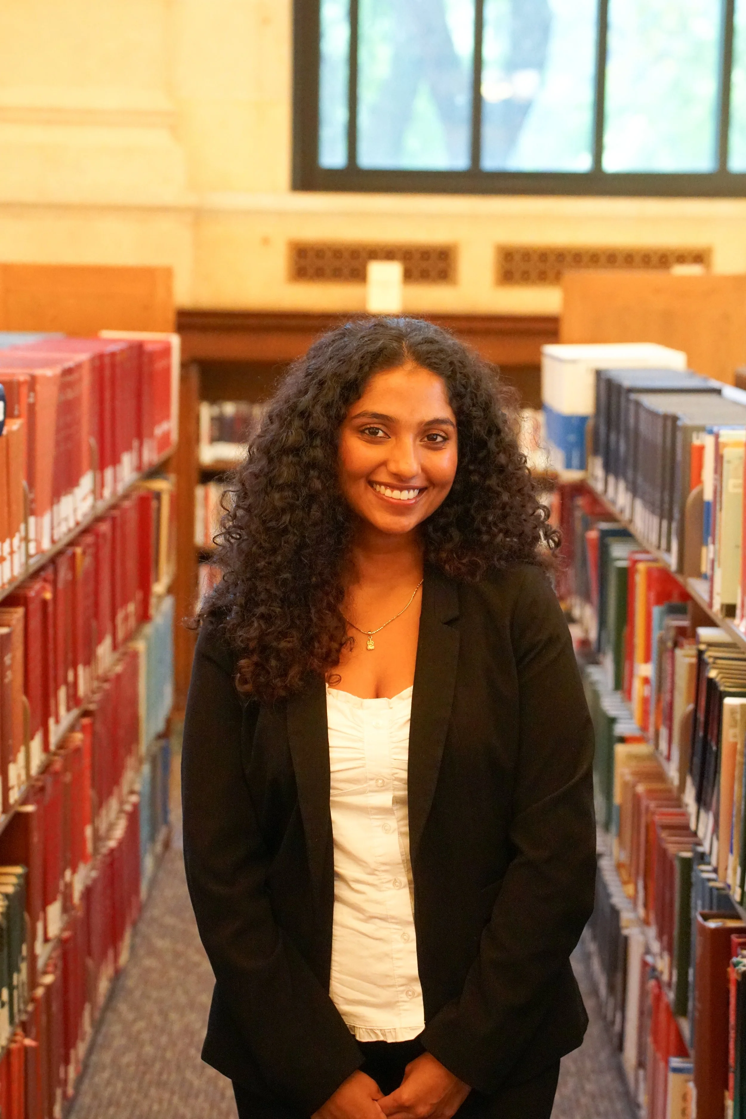A woman with curly hair, wearing a black blazer over a white top, stands on outdoor stairs in front of a building with large wooden doors and stone columns, smiling at the camera.