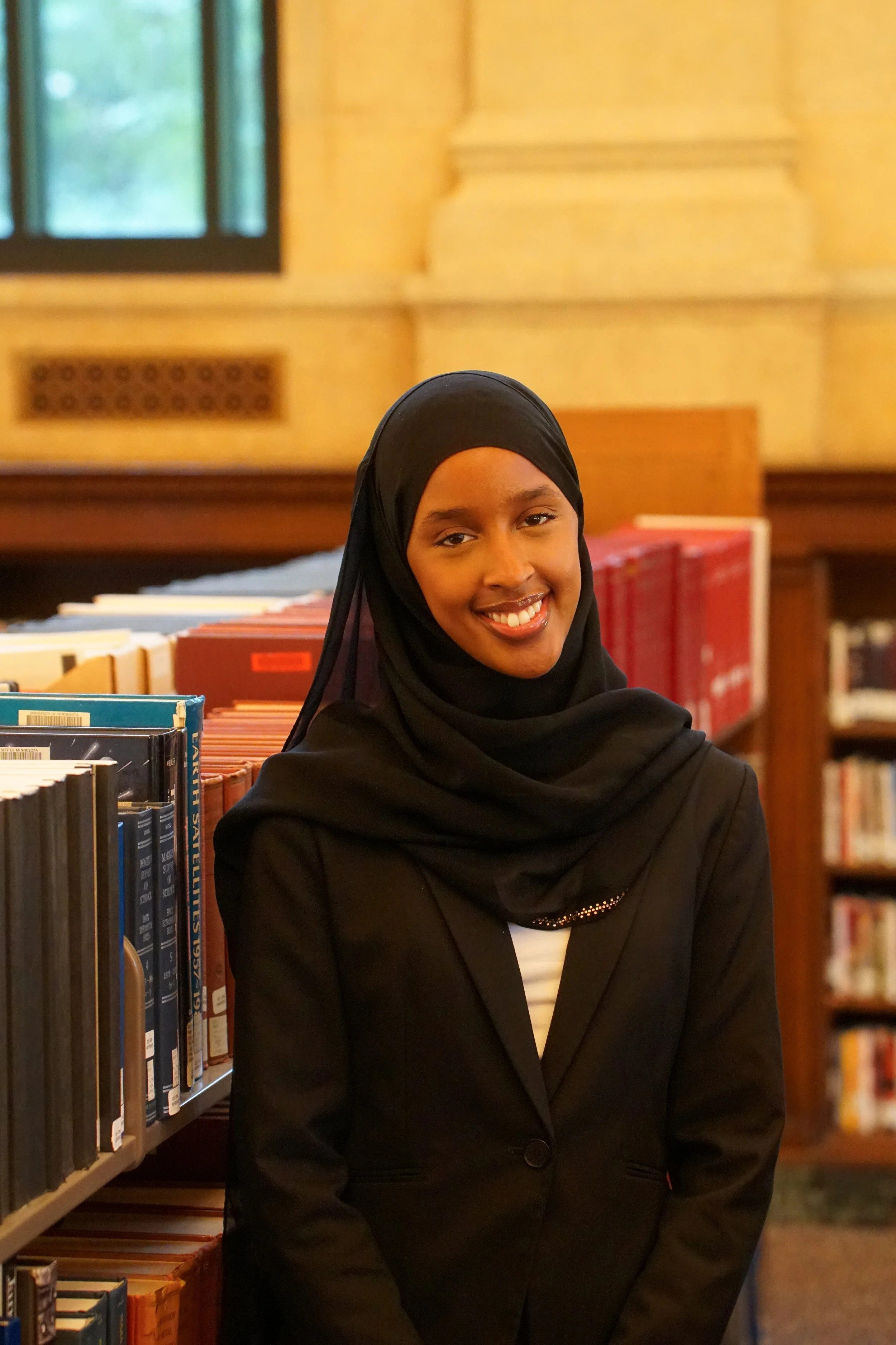 A woman wearing a black outfit and patterned headscarf standing on the steps of a building with wooden doors and stone columns.