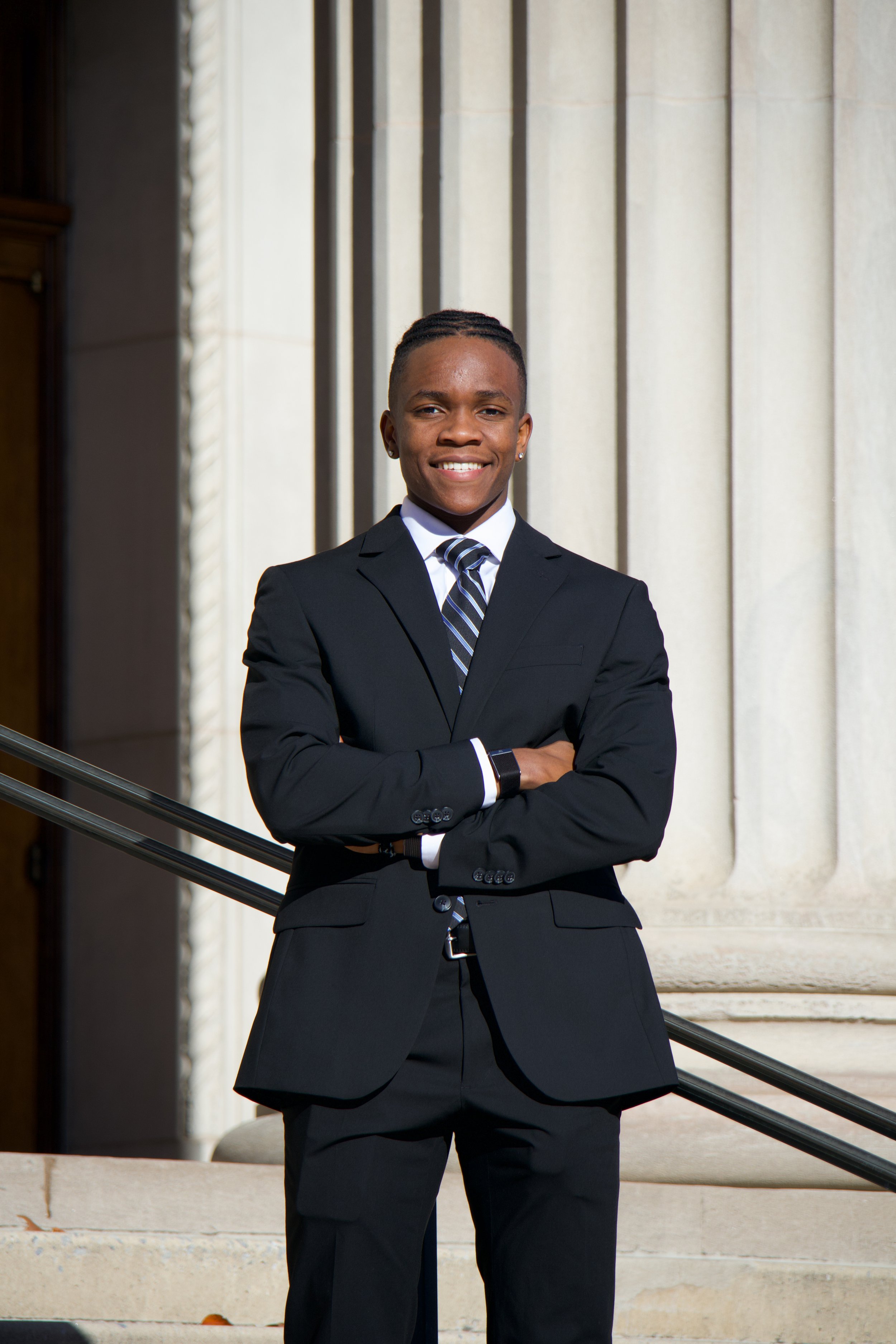 Young man wearing a suit and tie, standing confidently with arms crossed in front of a grand stone building with columns.