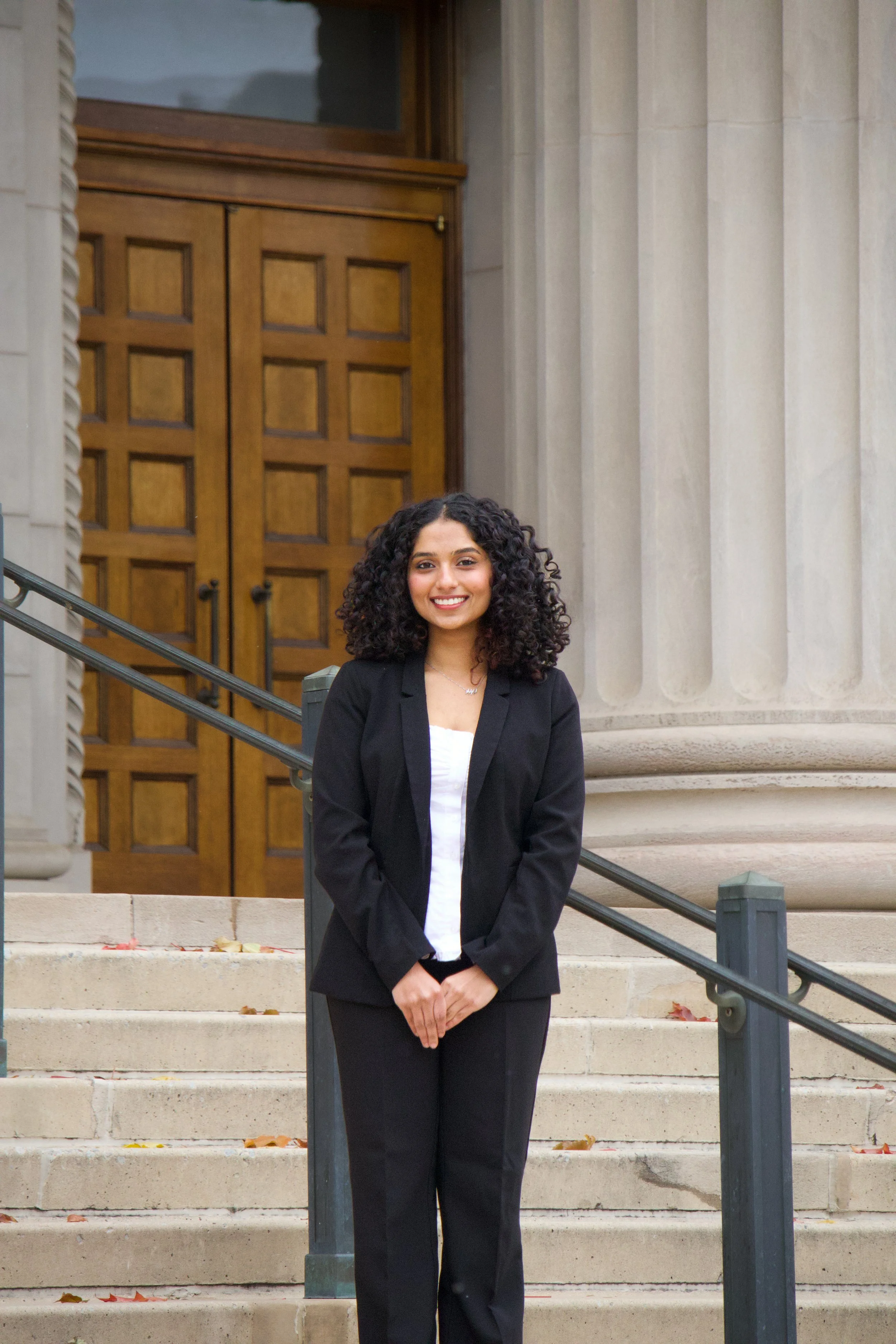 A woman with curly hair, wearing a black blazer over a white top, stands on outdoor stairs in front of a building with large wooden doors and stone columns, smiling at the camera.