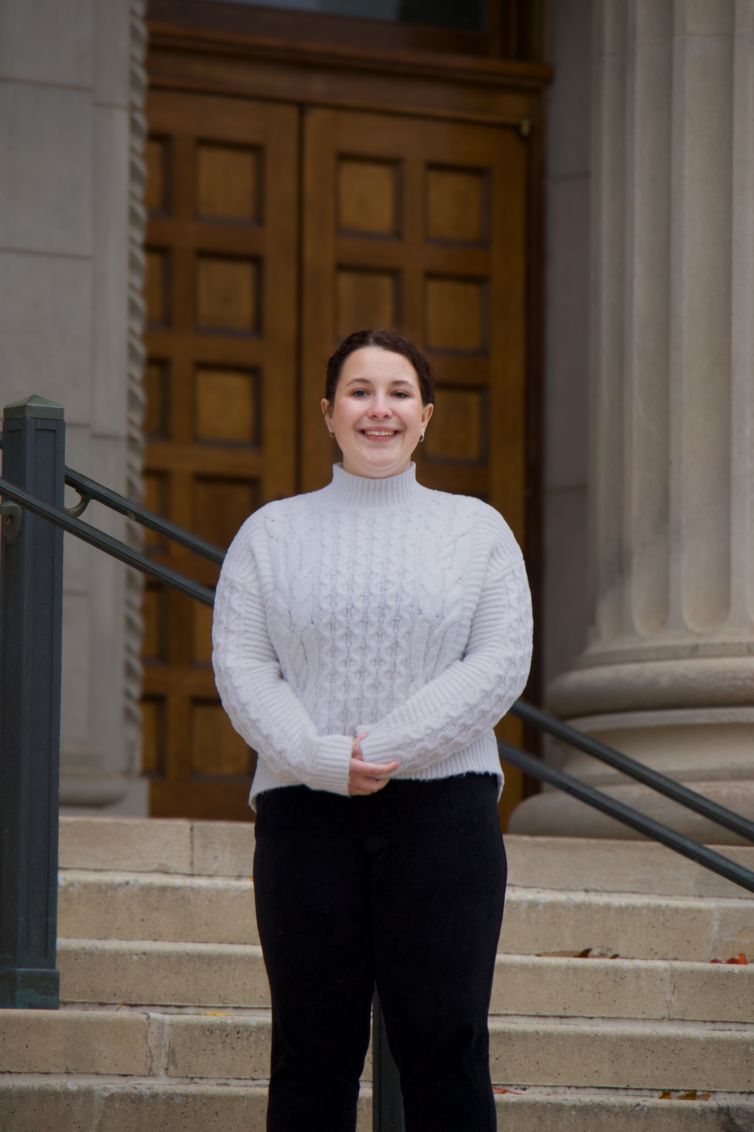 A woman standing on steps in front of a large wooden door with columns and stone walls, smiling and wearing a white sweater and black skirt.