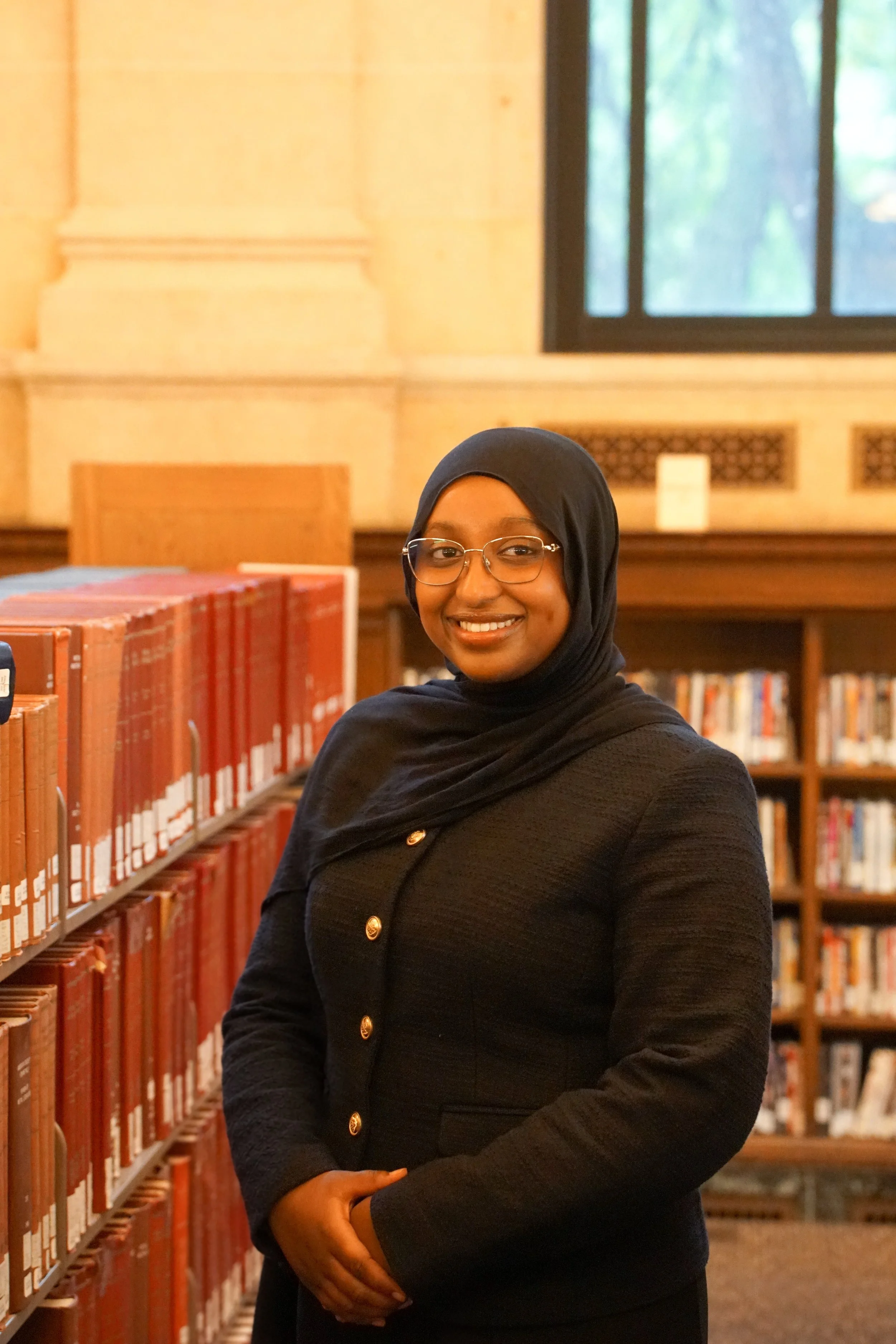 A woman wearing a black hijab, glasses, a white long sleeve top, and a black skirt, smiling and standing outdoors near stairs with a railing.