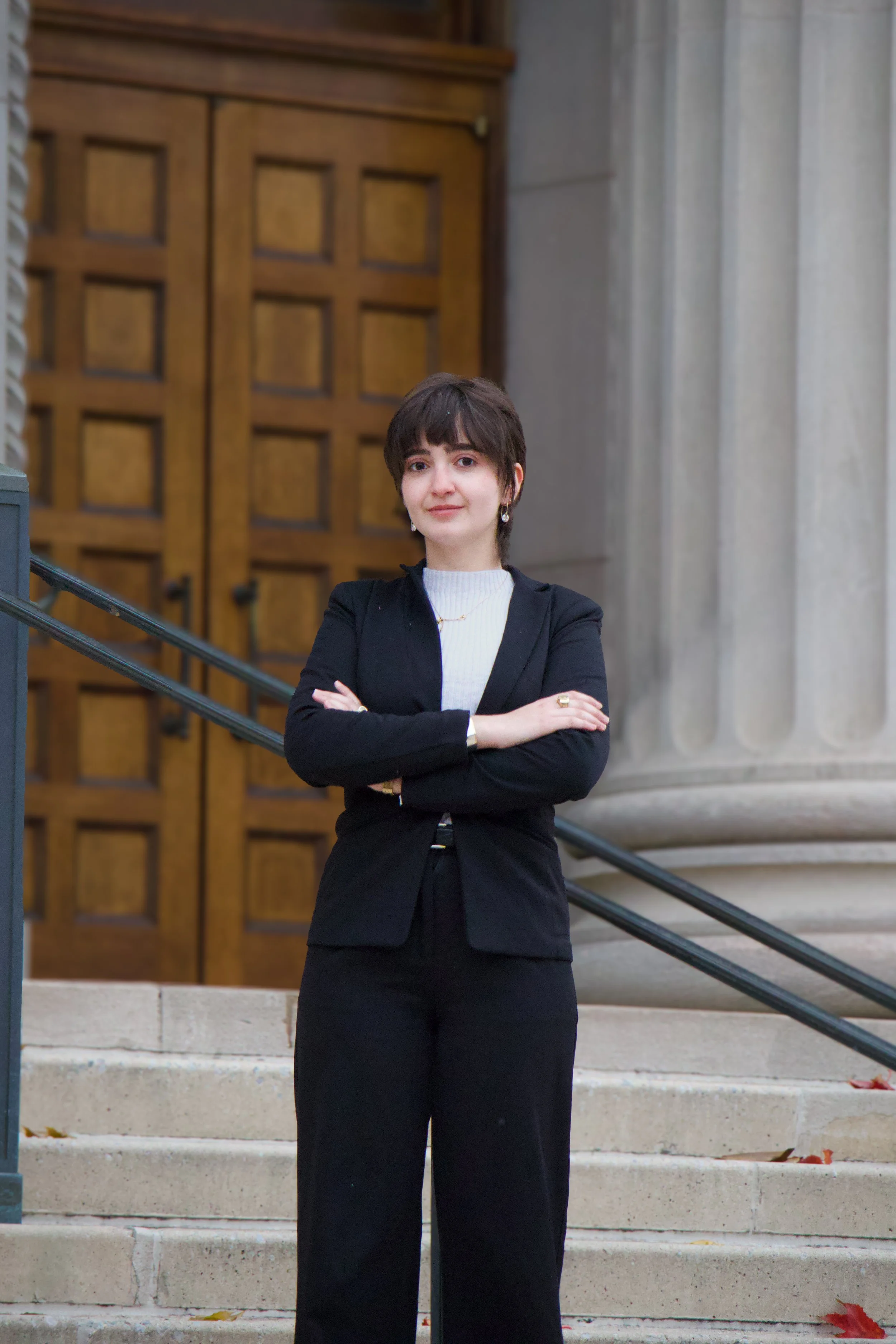 Portrait of a woman standing on stone steps in front of a large wooden door, wearing a black suit with arms crossed and looking at the camera.