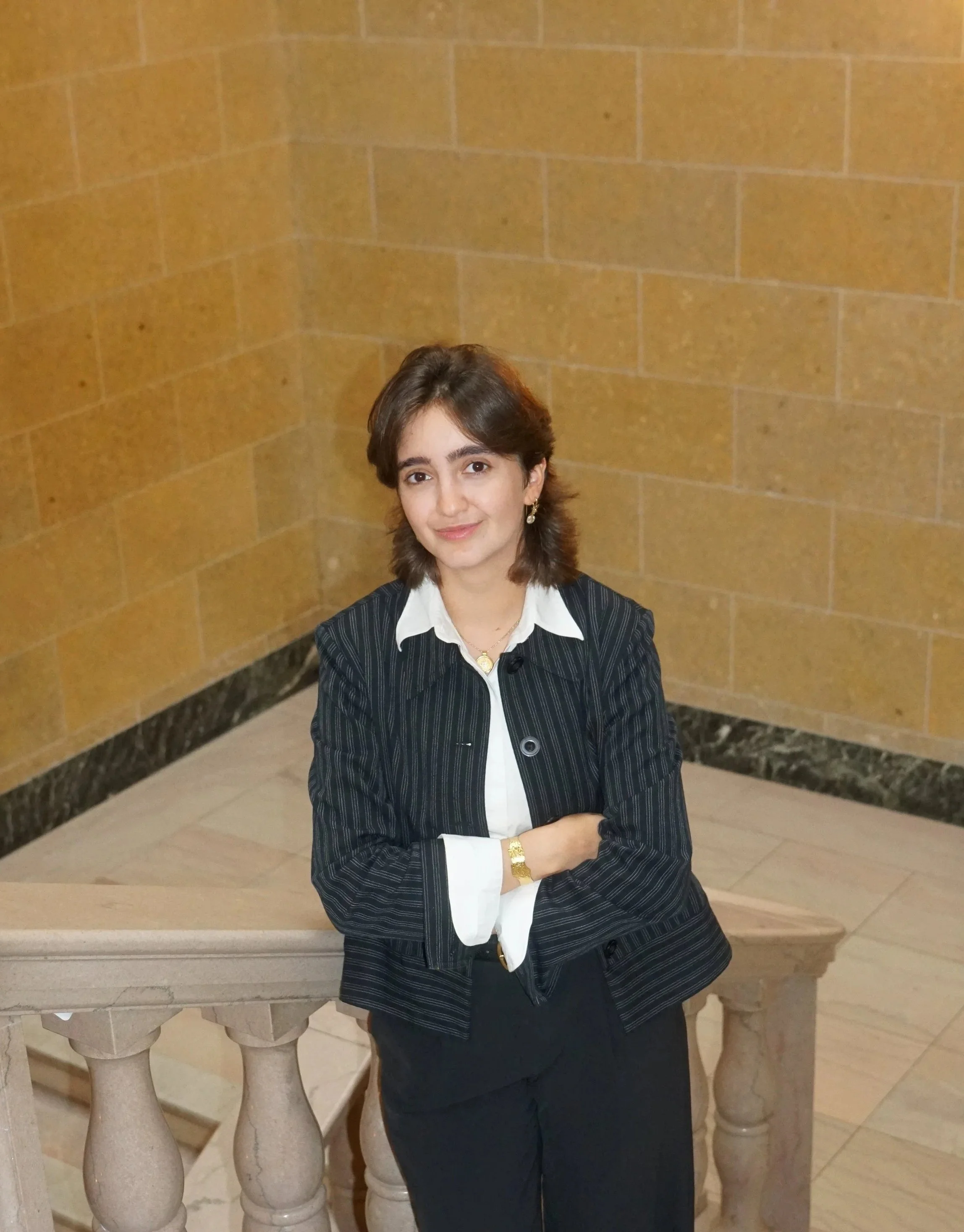 Portrait of a woman standing on stone steps in front of a large wooden door, wearing a black suit with arms crossed and looking at the camera.