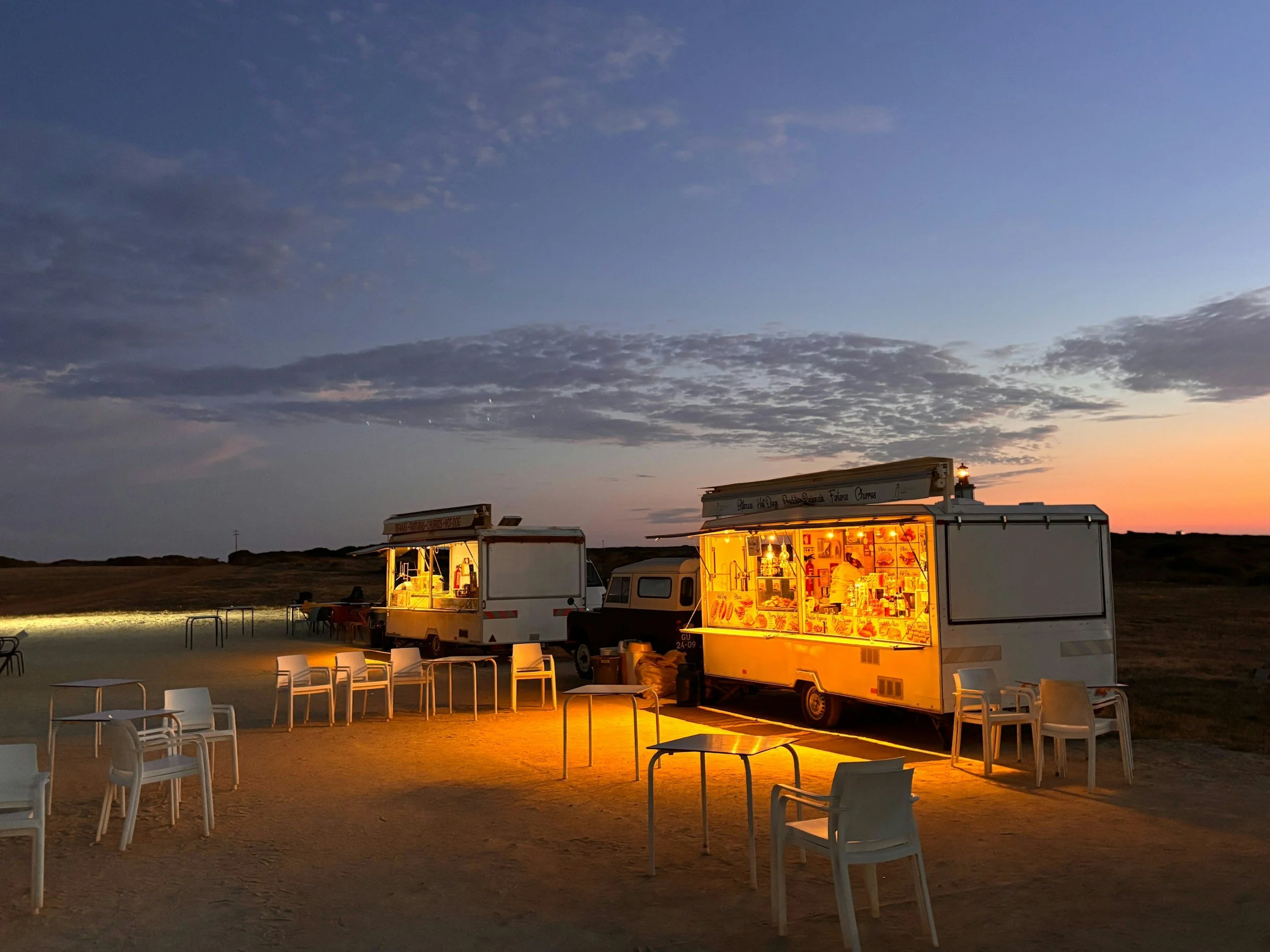 Food trucks illuminated with warm yellow lights at dusk, with a sky of scattered clouds and fading sunset in the background. White chairs and tables set up outside for seating.