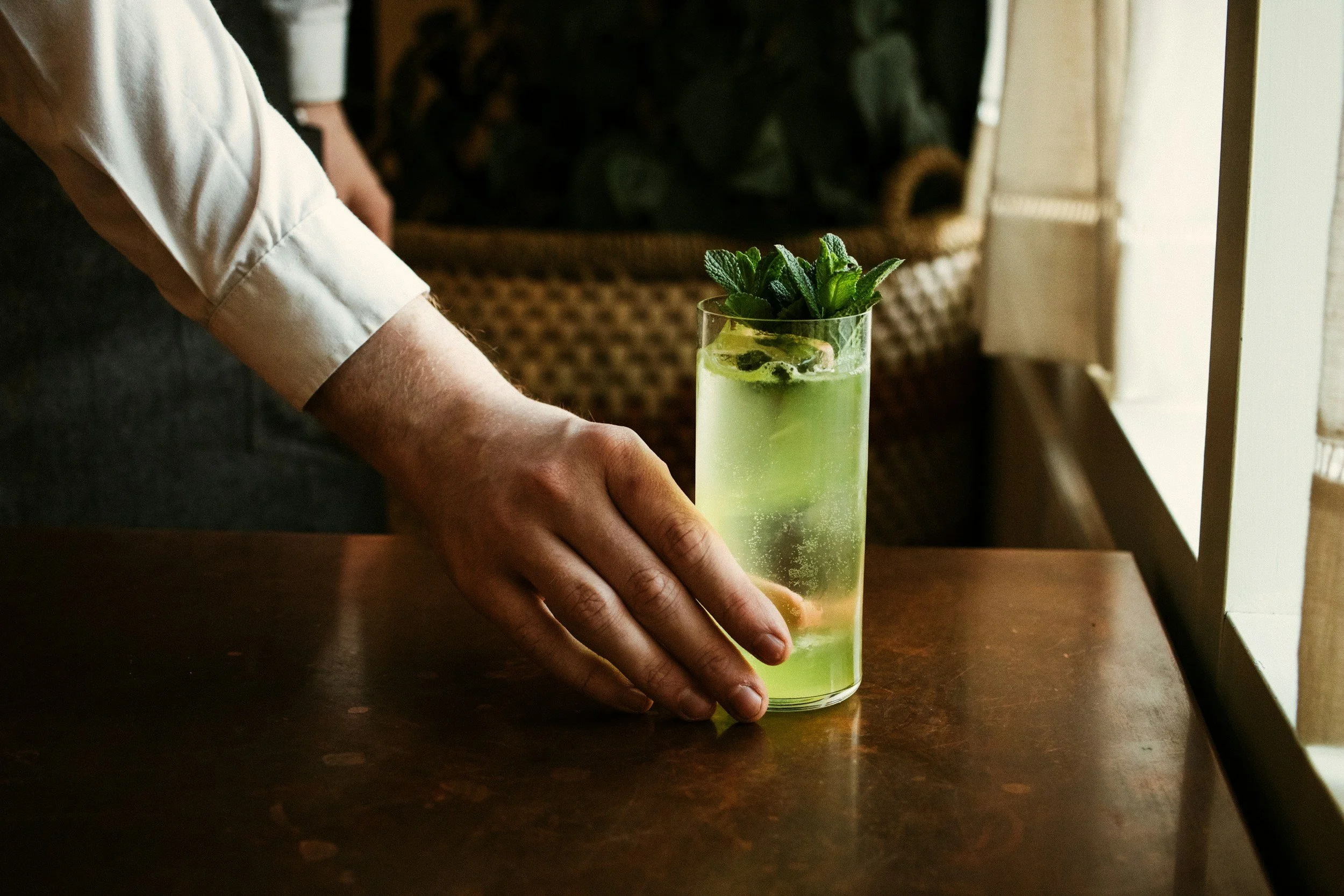 A person wearing a white shirt places a hand on a dark wooden table next to a tall glass of light green beverage garnished with fresh mint leaves