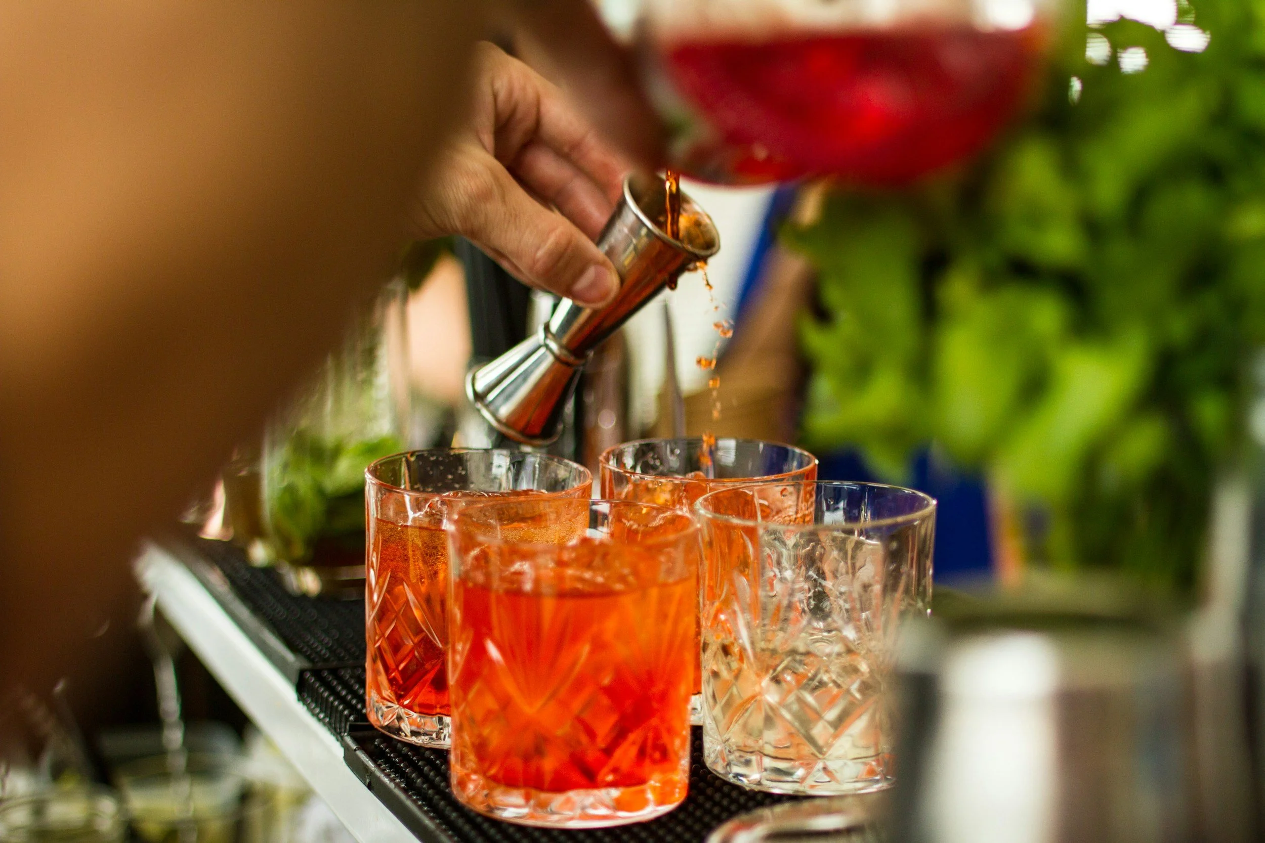 Close-up of an orange cocktail in a textured glass being filled from a chiller with a metal jigger. Multiple similar glasses are arranged on a black bar shelf.