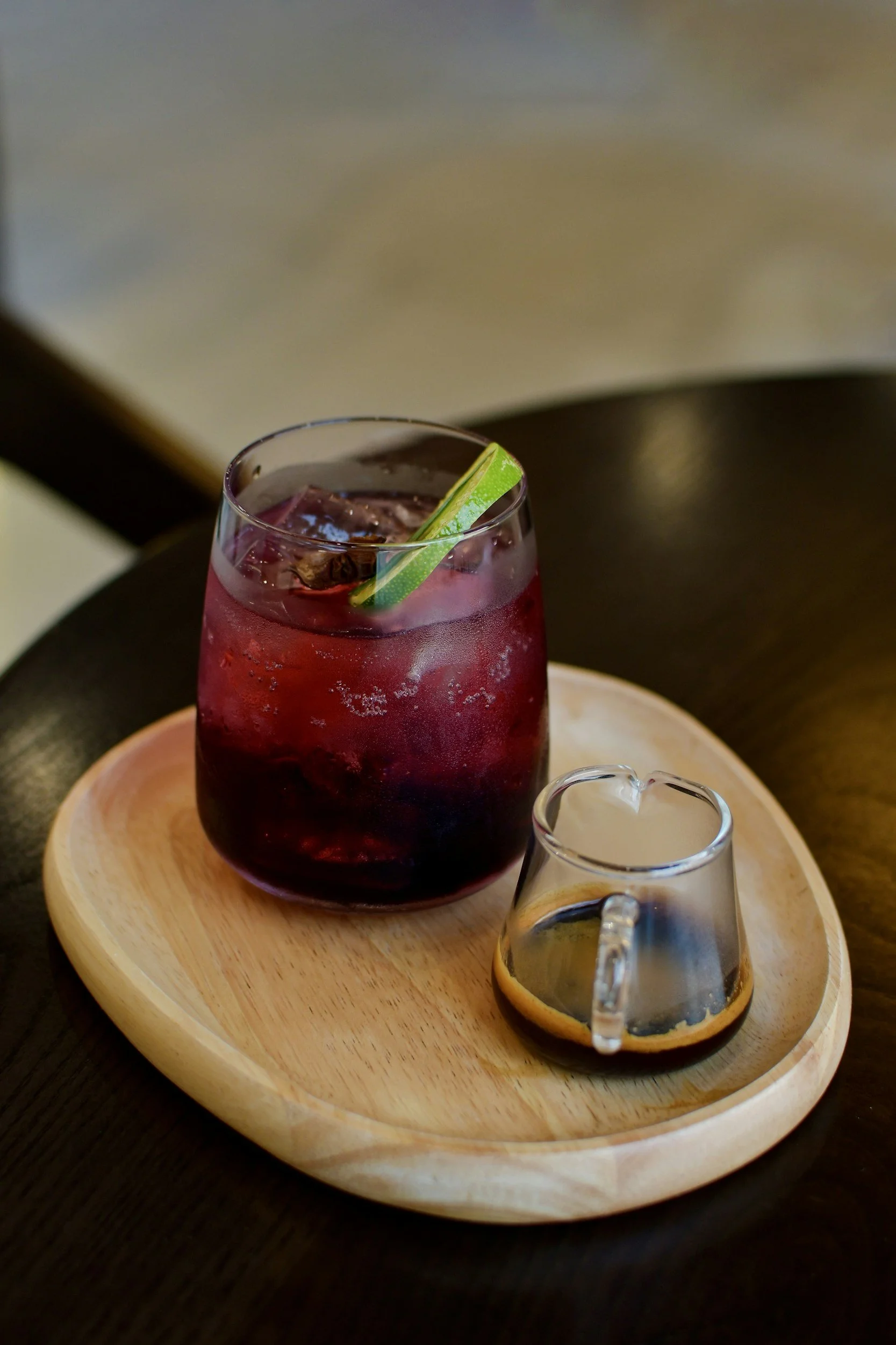 A glass of red iced beverage with a lime wedge garnish, placed on a wooden tray alongside a small glass pitcher of dark syrup or sauce.