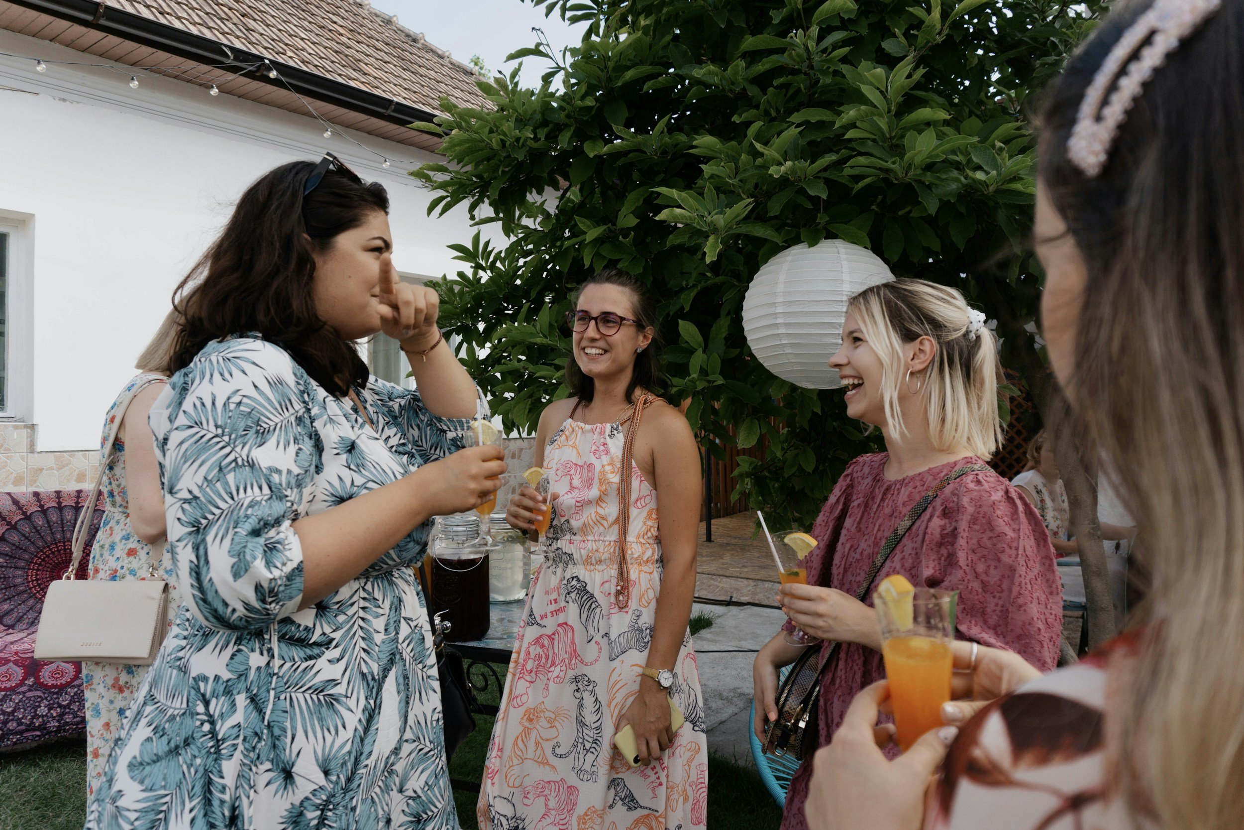 A group of women socializing outdoors, holding drinks with lemon slices, in a backyard with a house, greenery, and hanging paper lanterns.