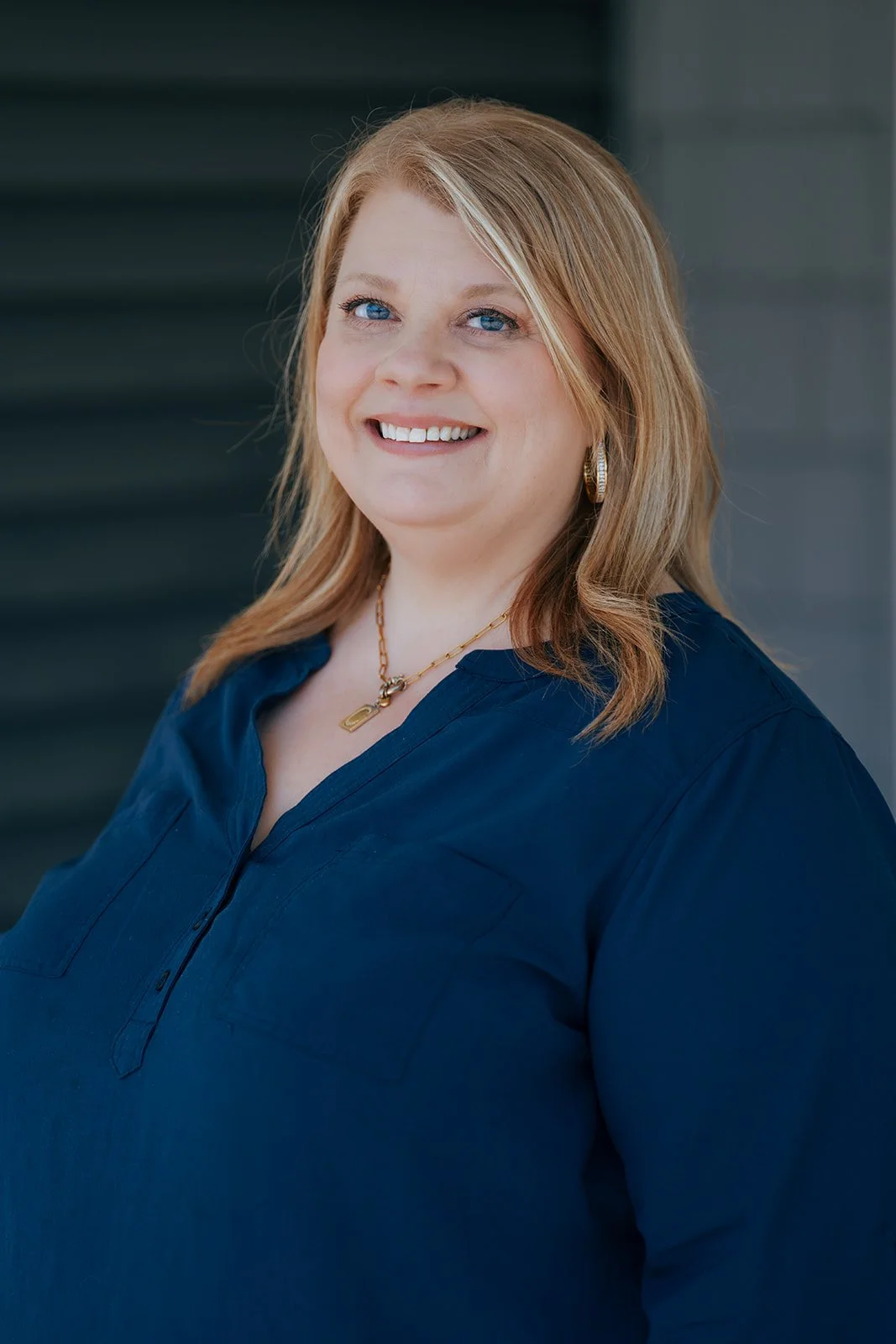 A woman with blonde hair, blue eyes, and a friendly smile, wearing a navy blue blouse, gold jewelry, and earrings, standing outdoors at Avielle Haven in Midland, Michigan.