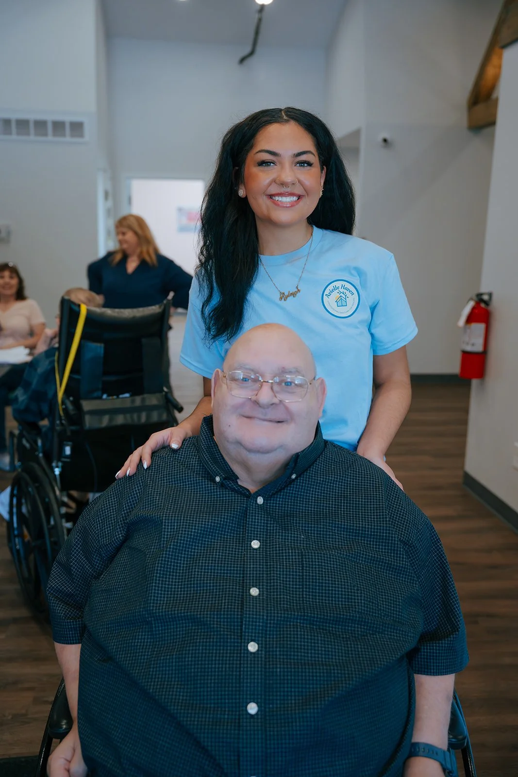 A young woman caregiver with dark hair smiling and standing behind a smiling elderly man with glasses in a wheelchair in Avielle Haven in Midland, Michigan.