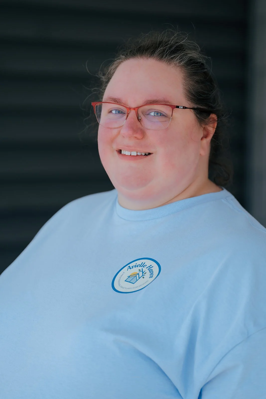 A smiling person with glasses wearing a light blue T-shirt with a logo that reads 'Avielle Haven'.