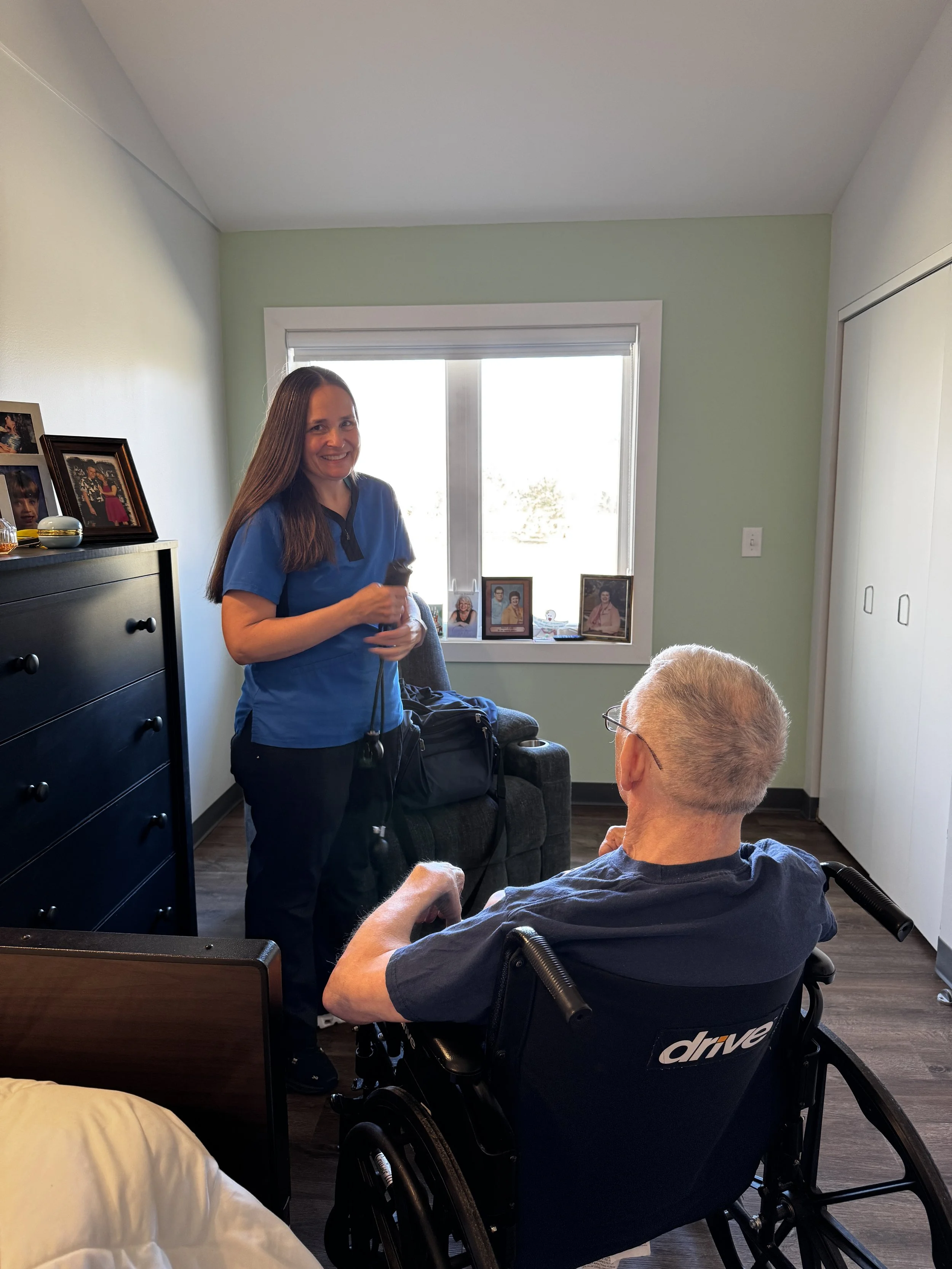 An RN in a blue uniform smiles at a man in a wheelchair. They are in a bright bedroom with a large window and framed photos. This reflects our long-term residential care, specialized memory care, and assisted living at Avielle Haven in Midland, MI