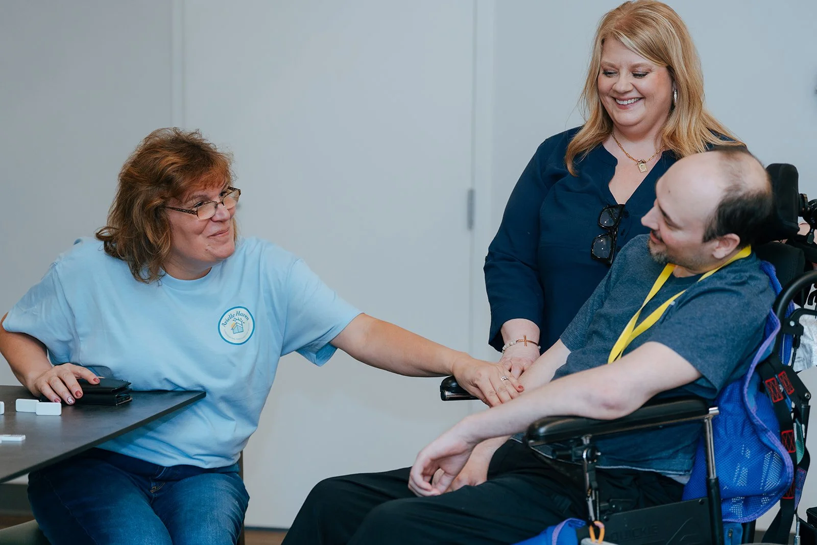 Two women hold hands with a smiling senior man in a wheelchair. One wears a white shirt and glasses. The blonde woman wears a blue shirt. They share a happy moment at Avielle Haven long-term residential care, specialized memory care, in Midland, MI.