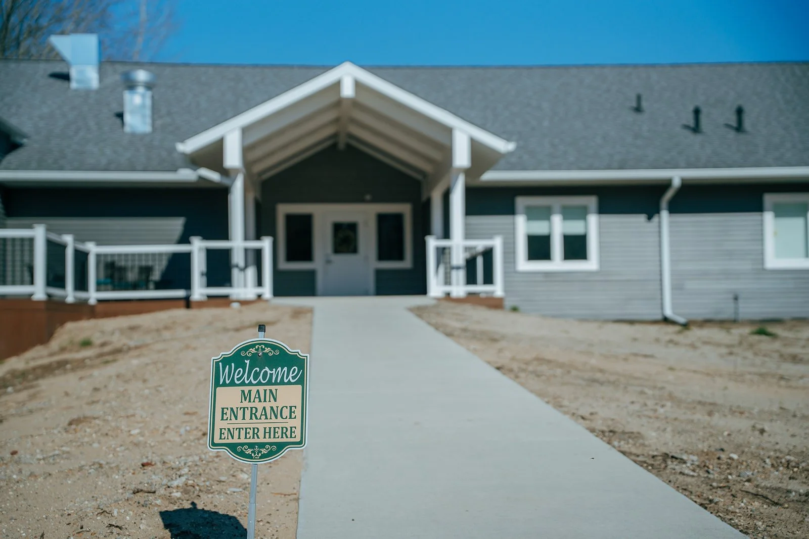 A modern house with a gray and white exterior, a front porch, and a sloped roof in Midland, Michigan. A sidewalk leads to the house, with a welcome sign indicating Avielle Haven main entrance.