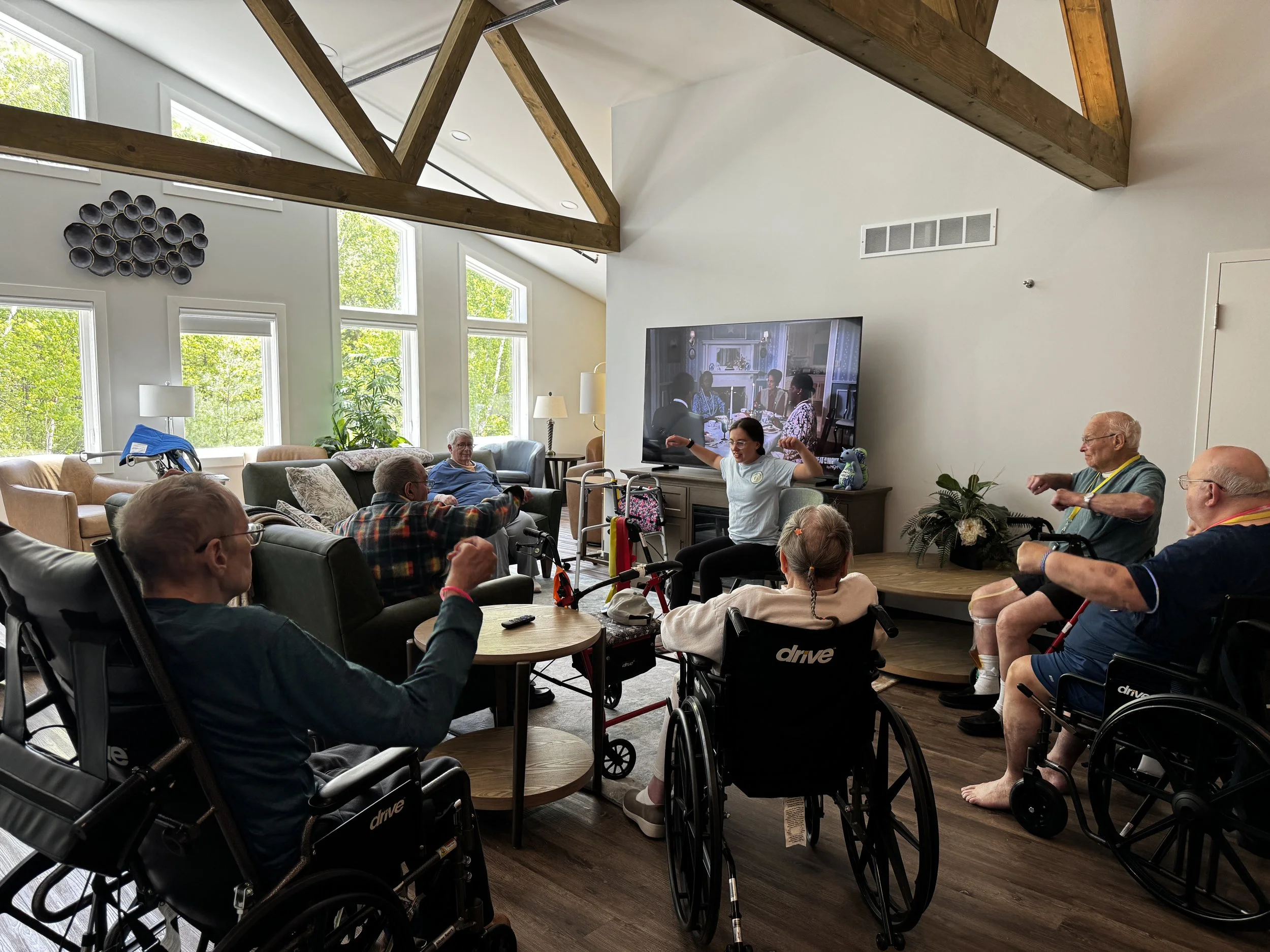 A group of elderly people, some in wheelchairs, watch television in a bright room with large windows and exposed wooden beams. Avielle Haven offers welcoming long-term residential care, specialized memory care, and assisted living Midland, MI.