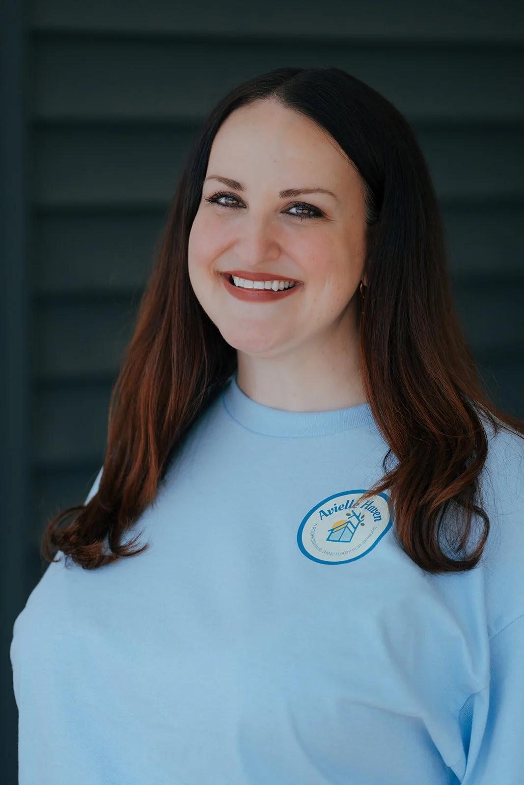 Smiling woman with brown hair wears a light blue Avielle Haven t-shirt with a house logo. She stands outdoors. She represents our assisted living, specialized memory care, and long-term residential care in Midland, MI.