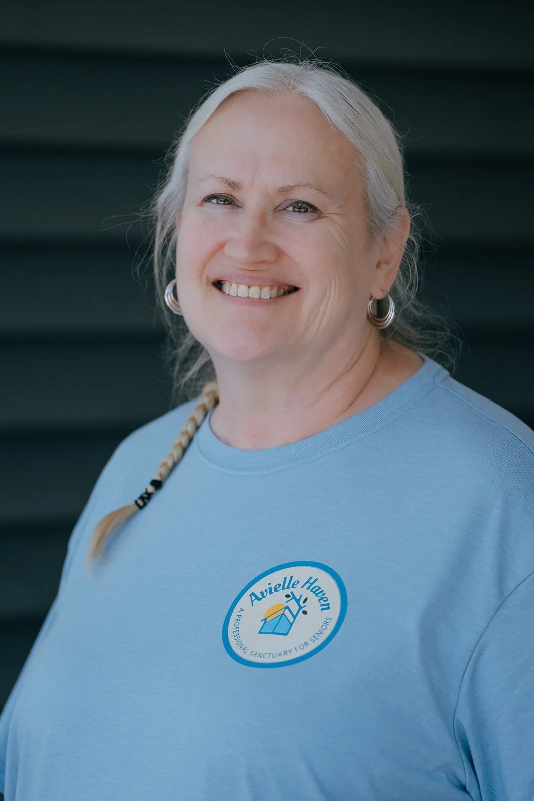 Smiling woman with gray braided hair wears a light blue Avielle Haven t-shirt against a dark background. She represents this sanctuary for seniors, offering long-term residential care, specialized memory care, and assisted living in Midland, MI