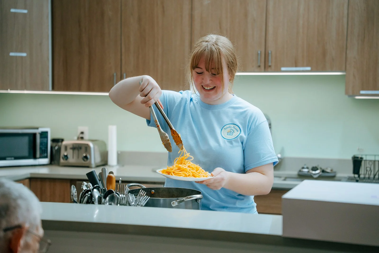 A woman serves spaghetti pasta with tongs in a kitchen. Residents enjoy home-cooked meals at Avielle Haven assisted living and memory care in Midland, MI.