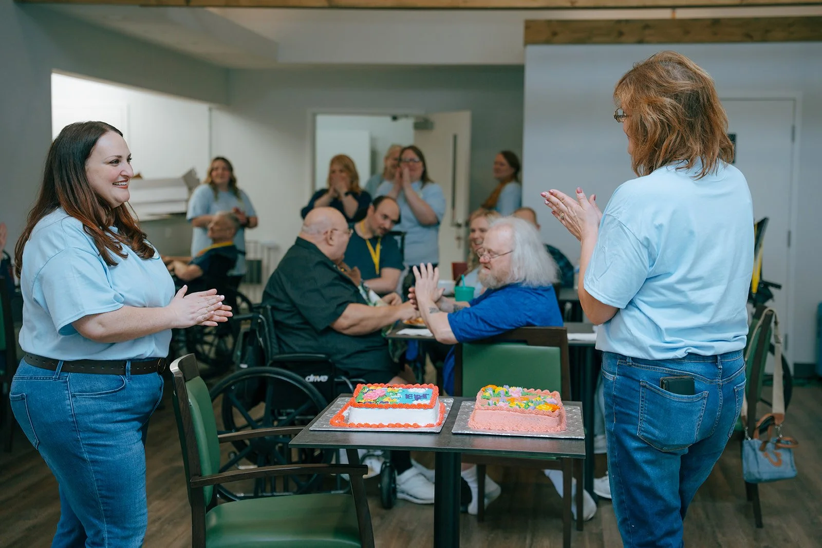 A joyful group celebrates around a table with two cakes. Two women smile and clap while residents in wheelchairs watch. They enjoy this social event at Avielle Haven assisted living and memory care in Midland, MI.