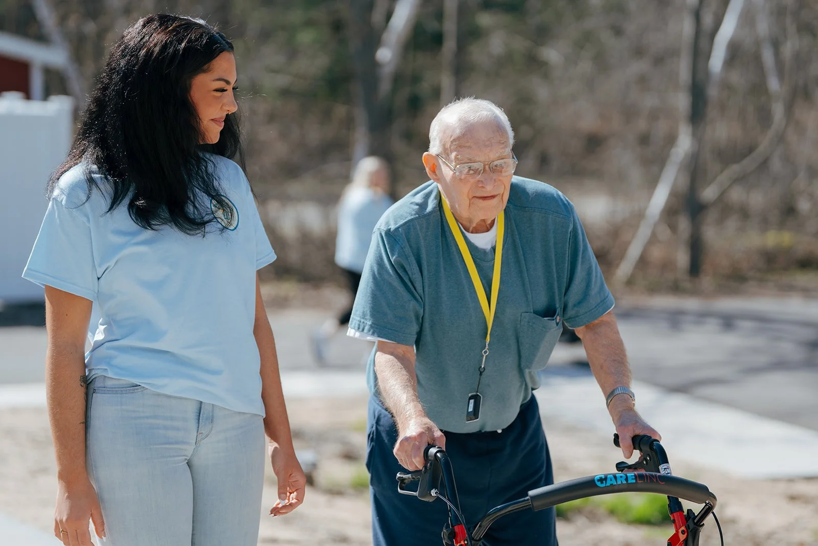 A young woman in light blue scrubs assists an elderly man using a walker outdoors on a sunny day. They enjoy the grounds at Avielle Haven, offering long-term residential care, specialized memory care, and assisted living in Midland, MI.