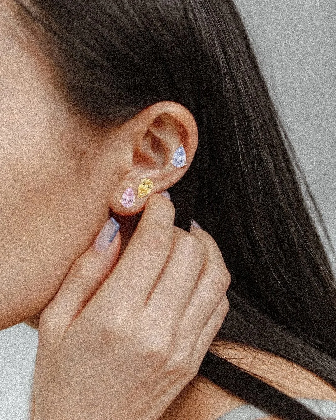 Close-up of a woman's ear with multiple colorful sapphire gemstone earrings, including pink, yellow, and blue stones, and her hand touching her hair.