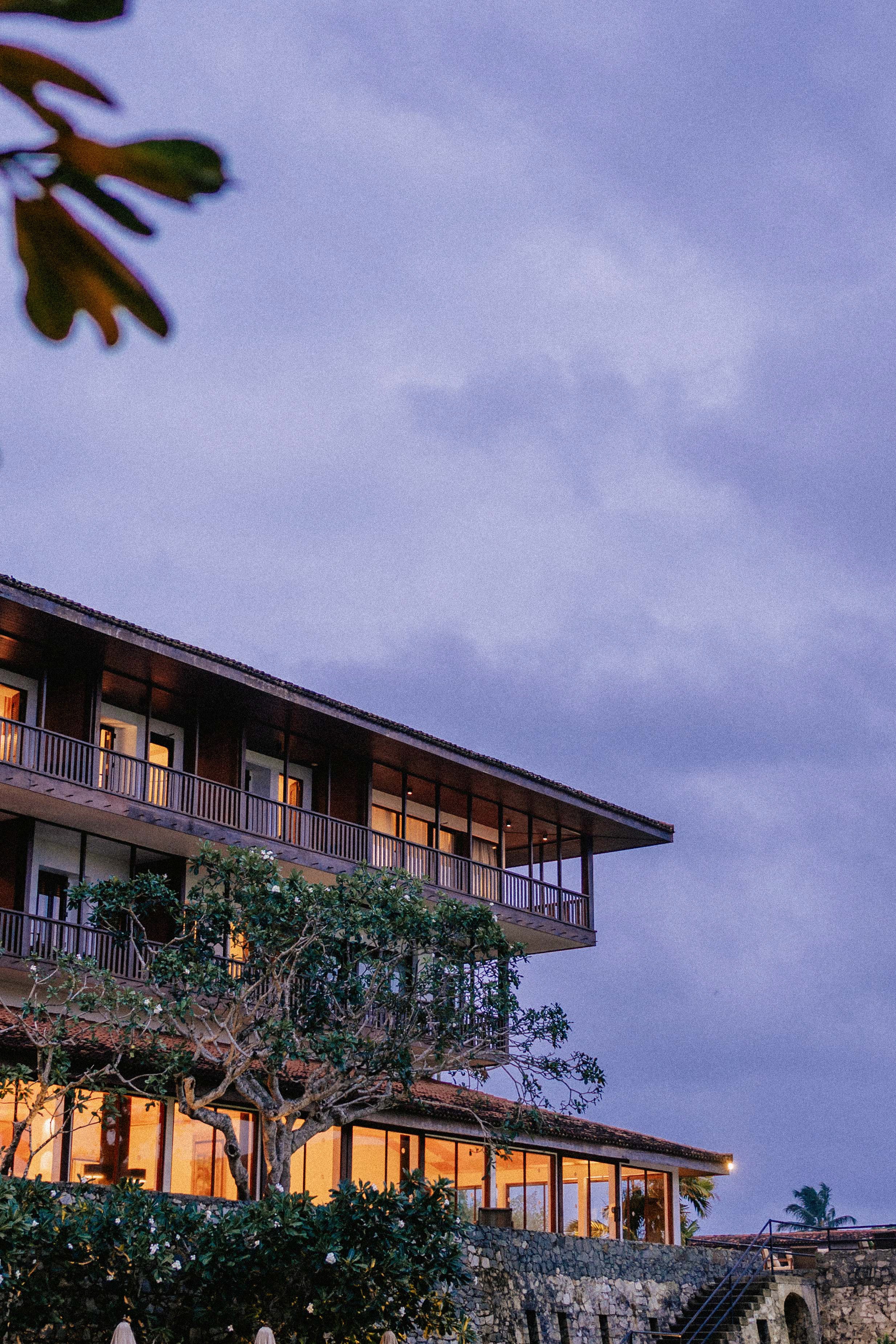 A multi-story building with large windows, trees with green leaves, and a stone wall, during dusk with a purple sky.