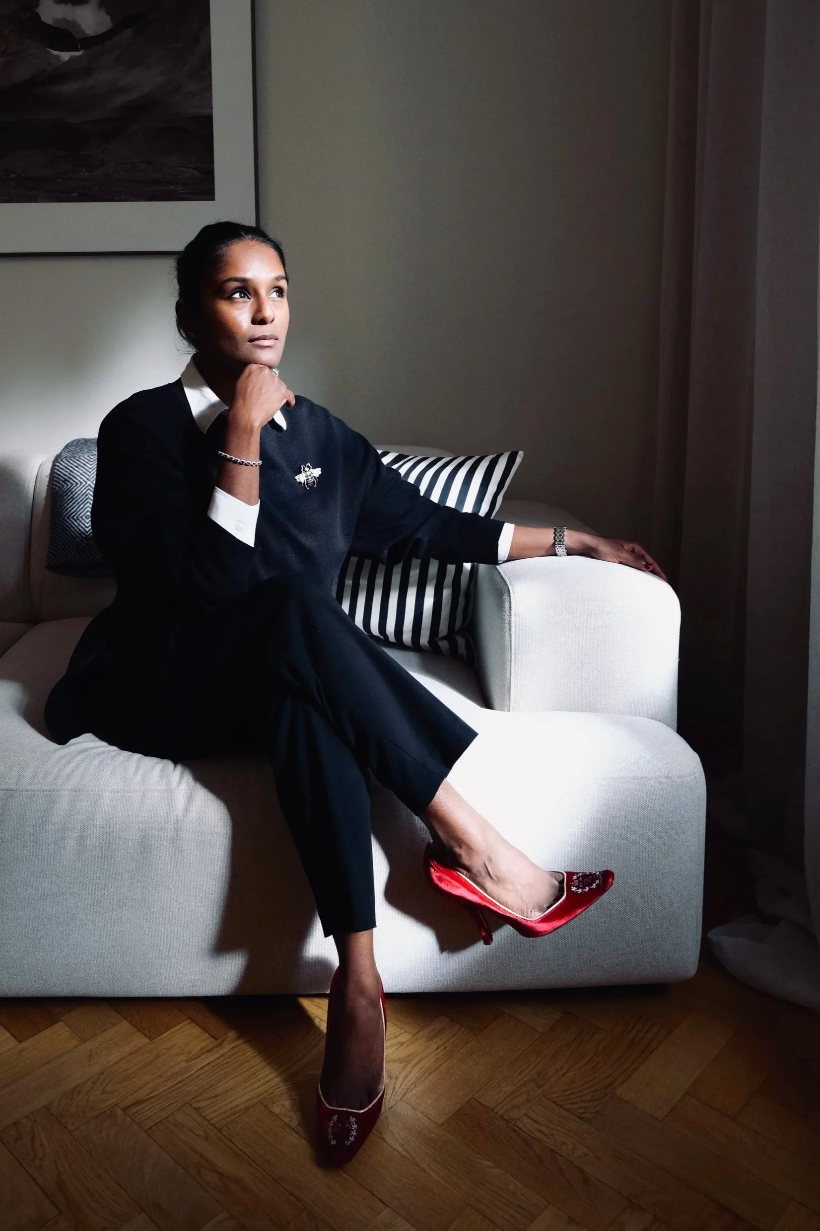 A woman sitting on a white couch, wearing a black sweater with white cuffs and collar, red high heels, and jewellery, looking thoughtfully to the side with her hand near her chin, in a dimly lit room with a painting on the wall and striped pillows.