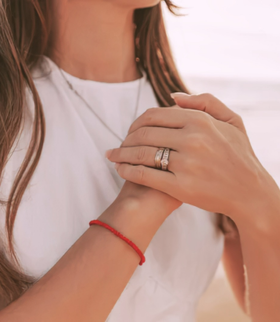 Mujer con manos juntas en una pose reflexiva, usando una cadena en el cuello, un anillo y una pulsera roja en la muñeca.