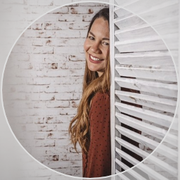 Mujer sonriendo mirando a través de una pared de lamas blancas en un entorno con pared de ladrillos blanca.