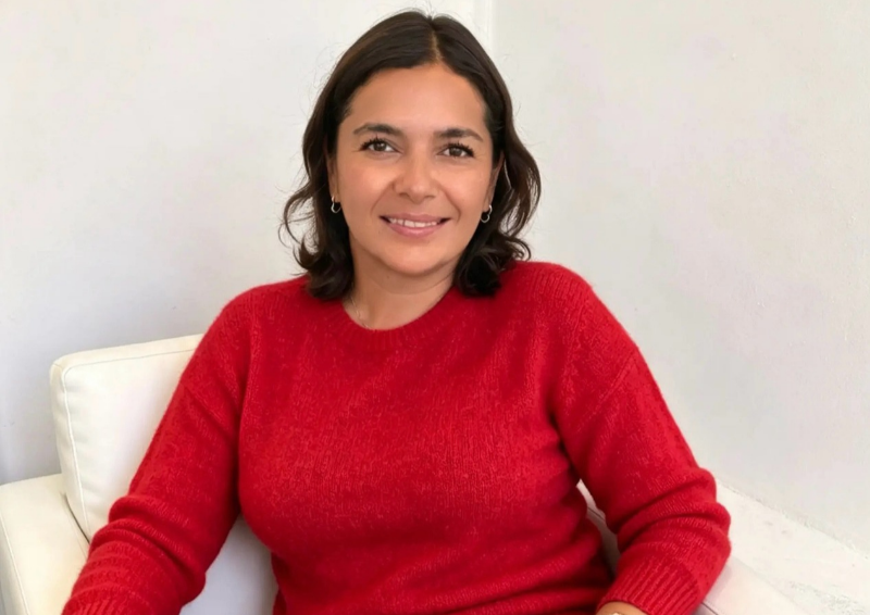 Mujer sonriendo con cabello castaño short y usando suéter rojo, sentada en un sillón blanco frente a una pared blanca.