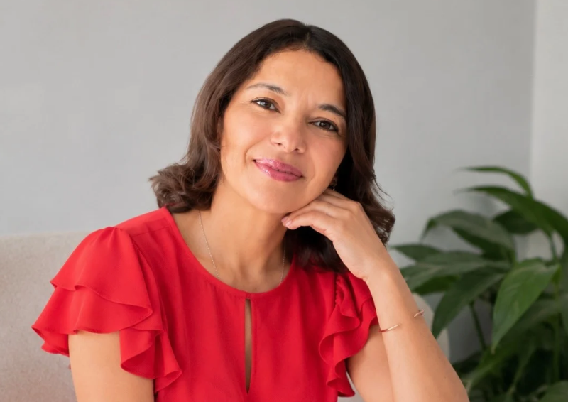 Mujer sonriendo, con cabello castaño, vestido rojo y fondo con plantas verdes en interior