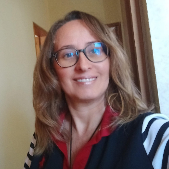 Mujer con gafas, cabello rubio y rizado, sonriendo, con camiseta de rayas negras y blancas, en un interior con ventana y pared beige.