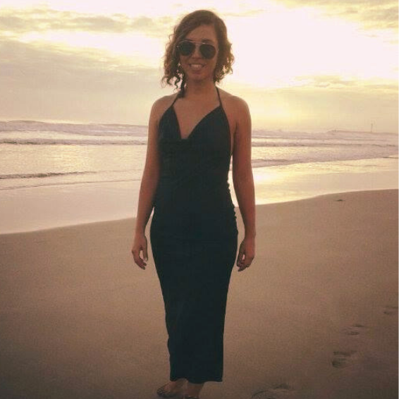 Mujer con vestido largo negro y gafas de sol en la playa al atardecer.
