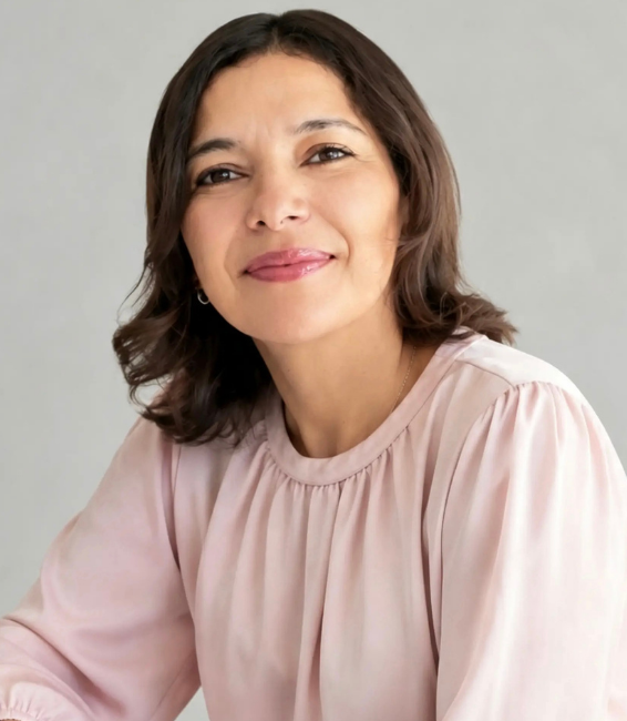 Retrato de una mujer con cabello castaño oscuro, sonriendo suavemente, usando blusa beige y pendientes pequeños, en un fondo gris claro.