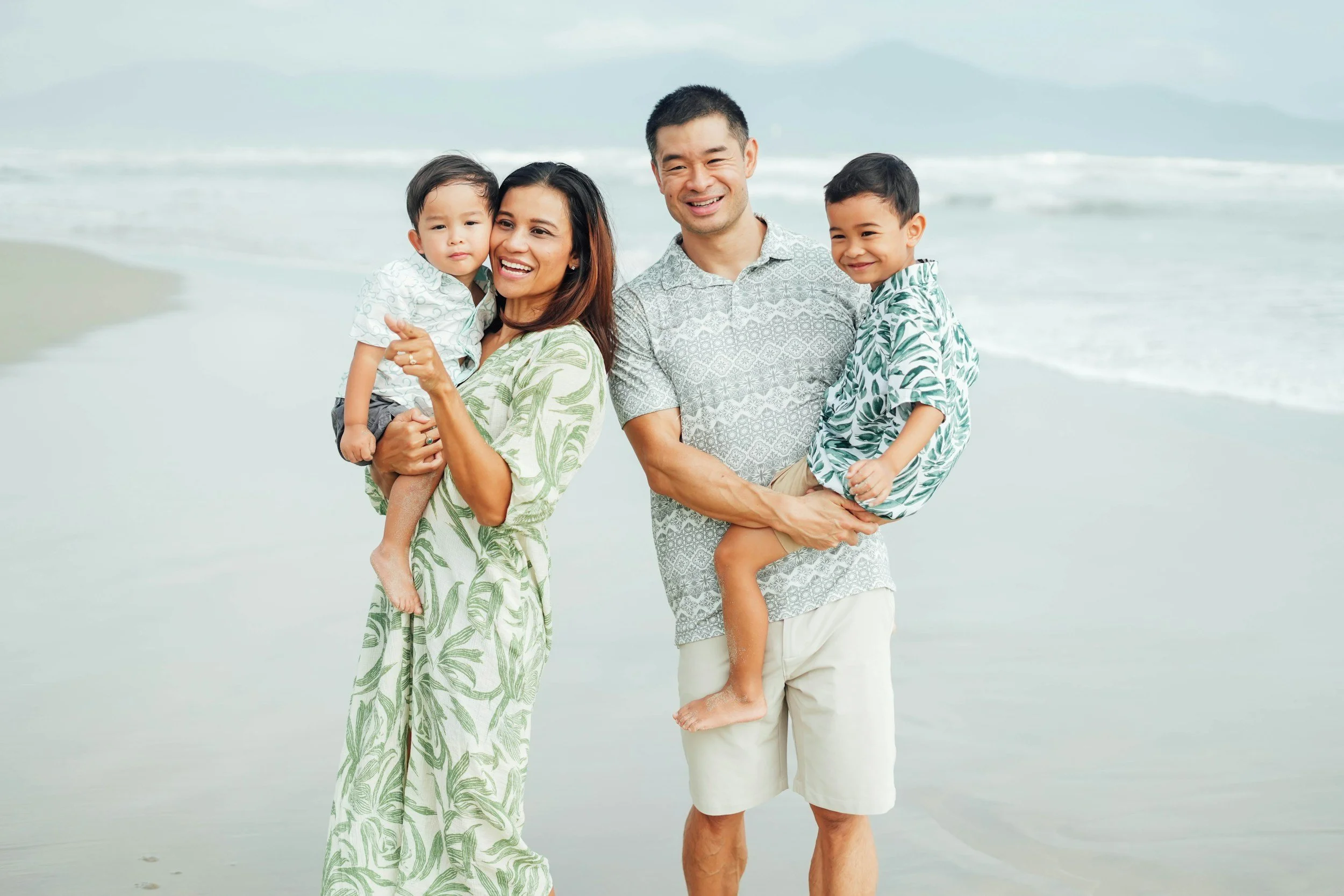 Family of four enjoying a day at the beach, with two children and two adults standing on the sand near the shoreline, smiling and dressed casually.