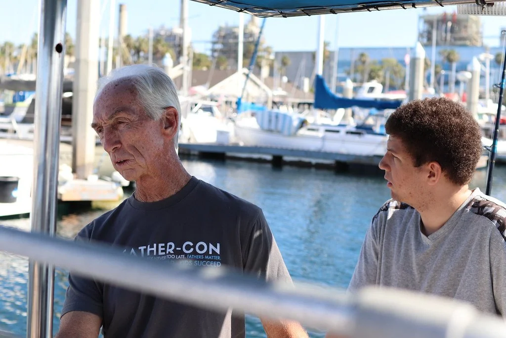 An elderly man and a young man having a conversation on a dock near boats at a marina.