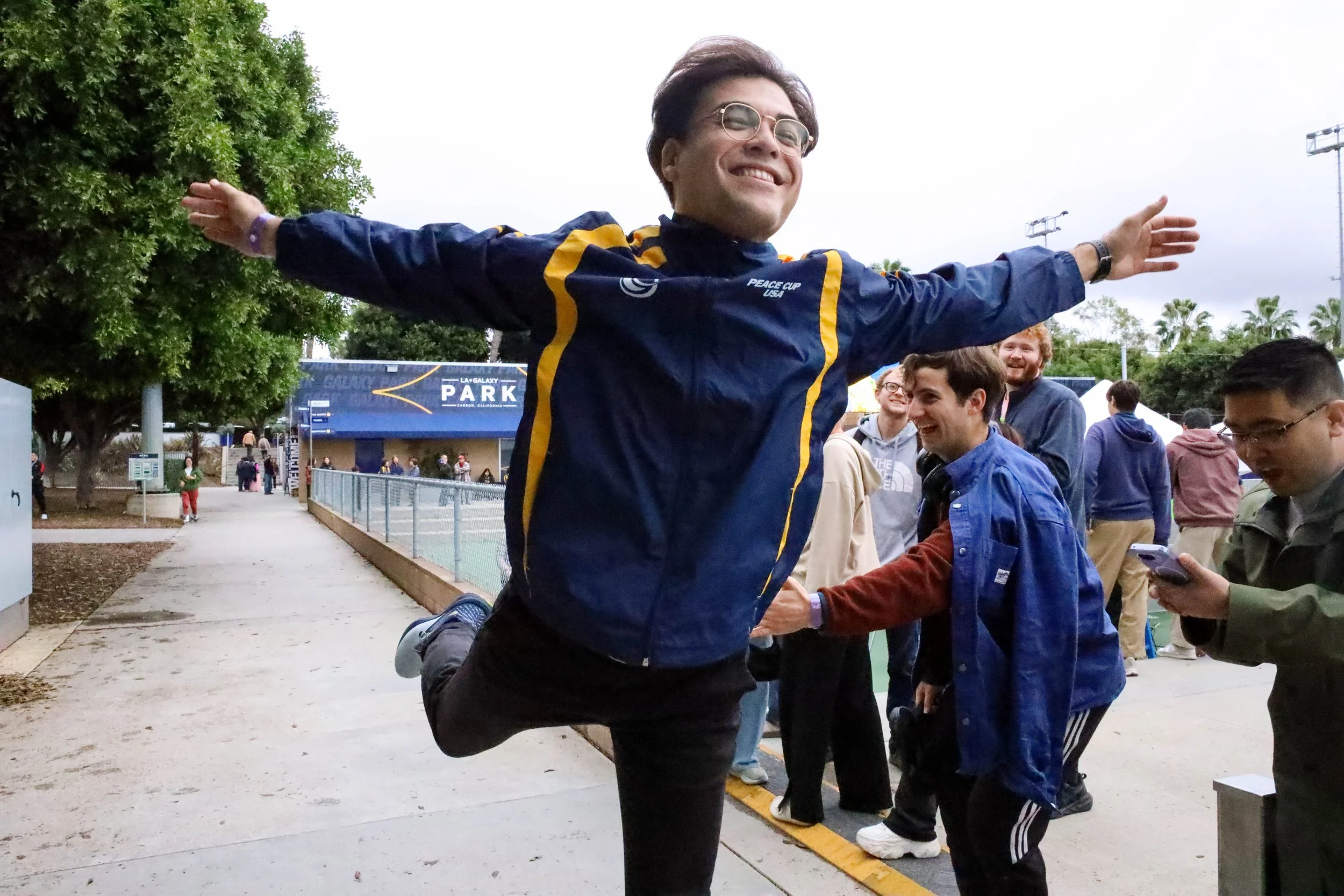 A person with glasses and short hair smiling with arms outstretched, standing in a group at an outdoor event near a sign for LA Galaxy Park in Carson, California.