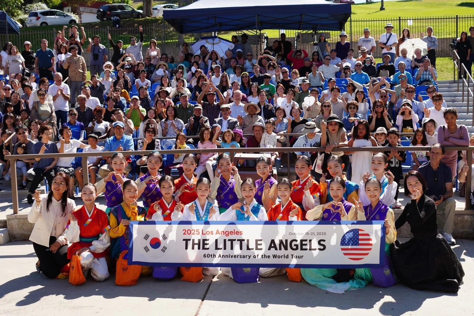 Group of children and adults gathered outdoors for a cultural performance or event, holding a banner that reads '2025 Los Angeles THE LITTLE ANGELS 60th Anniversary of the World Tour', featuring the flags of South Korea and the USA, with some individuals in traditional Korean hanbok costumes in the front row.