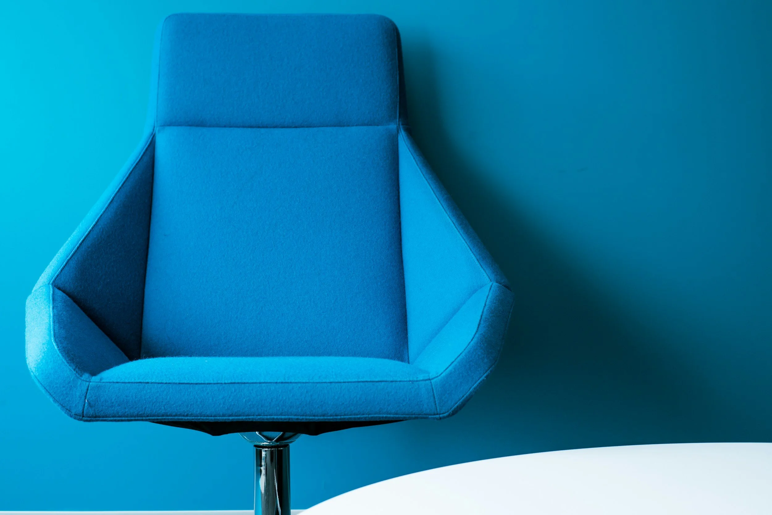 A blue office chair with a curved backrest and armrests, set against a matching blue wall and surface.