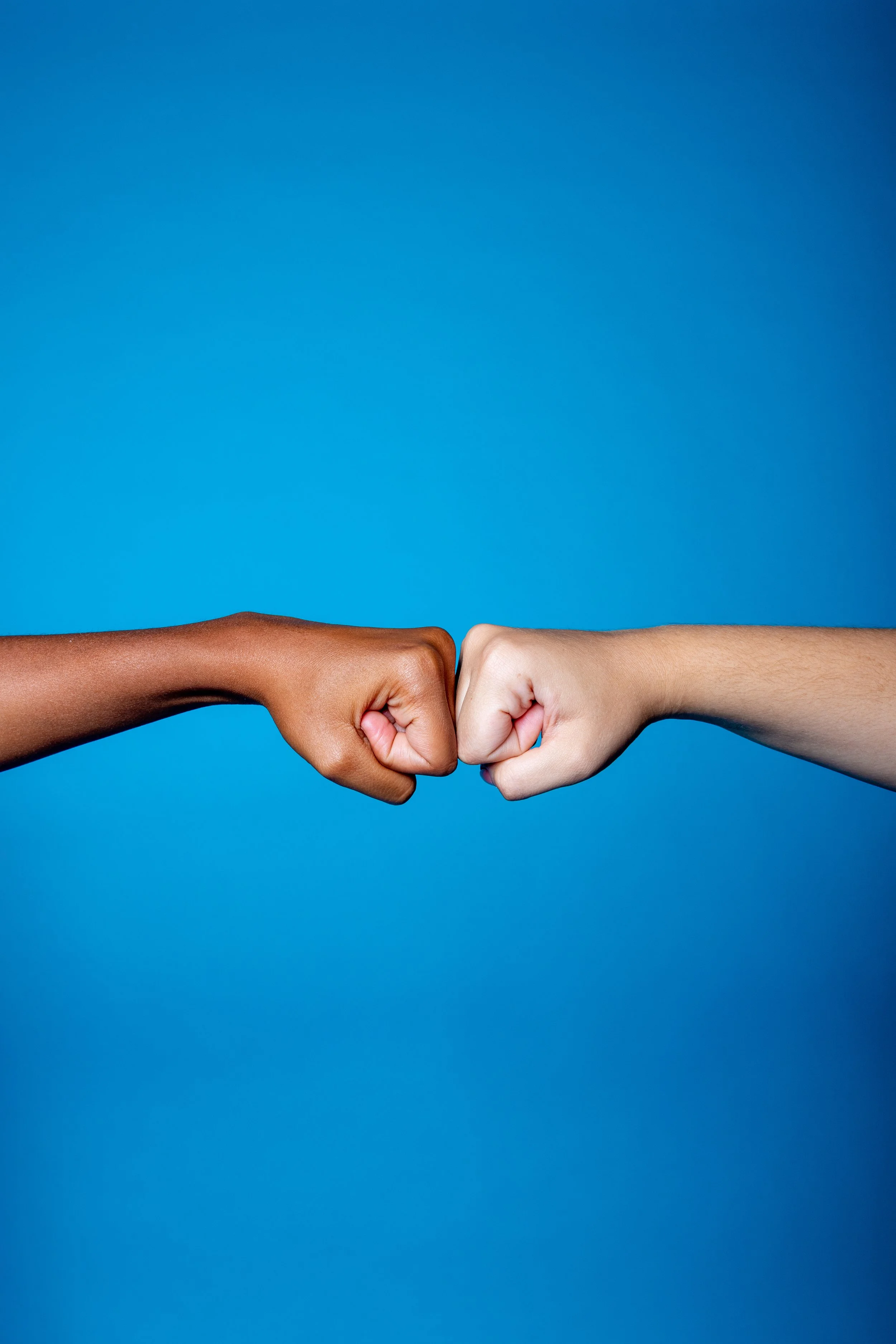 Two hands, one dark-skinned and one light-skinned, forming fists and touching at the knuckles against a blue background.