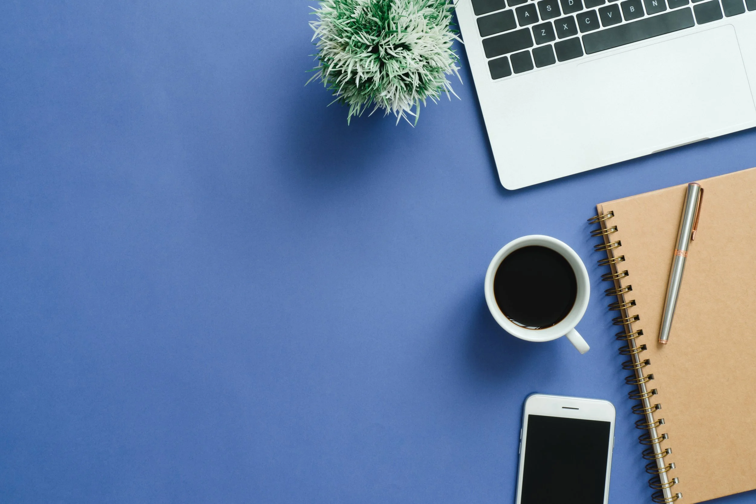 A laptop, a potted plant, a notebook with a pen, a cup of coffee, and a smartphone on a blue desk.