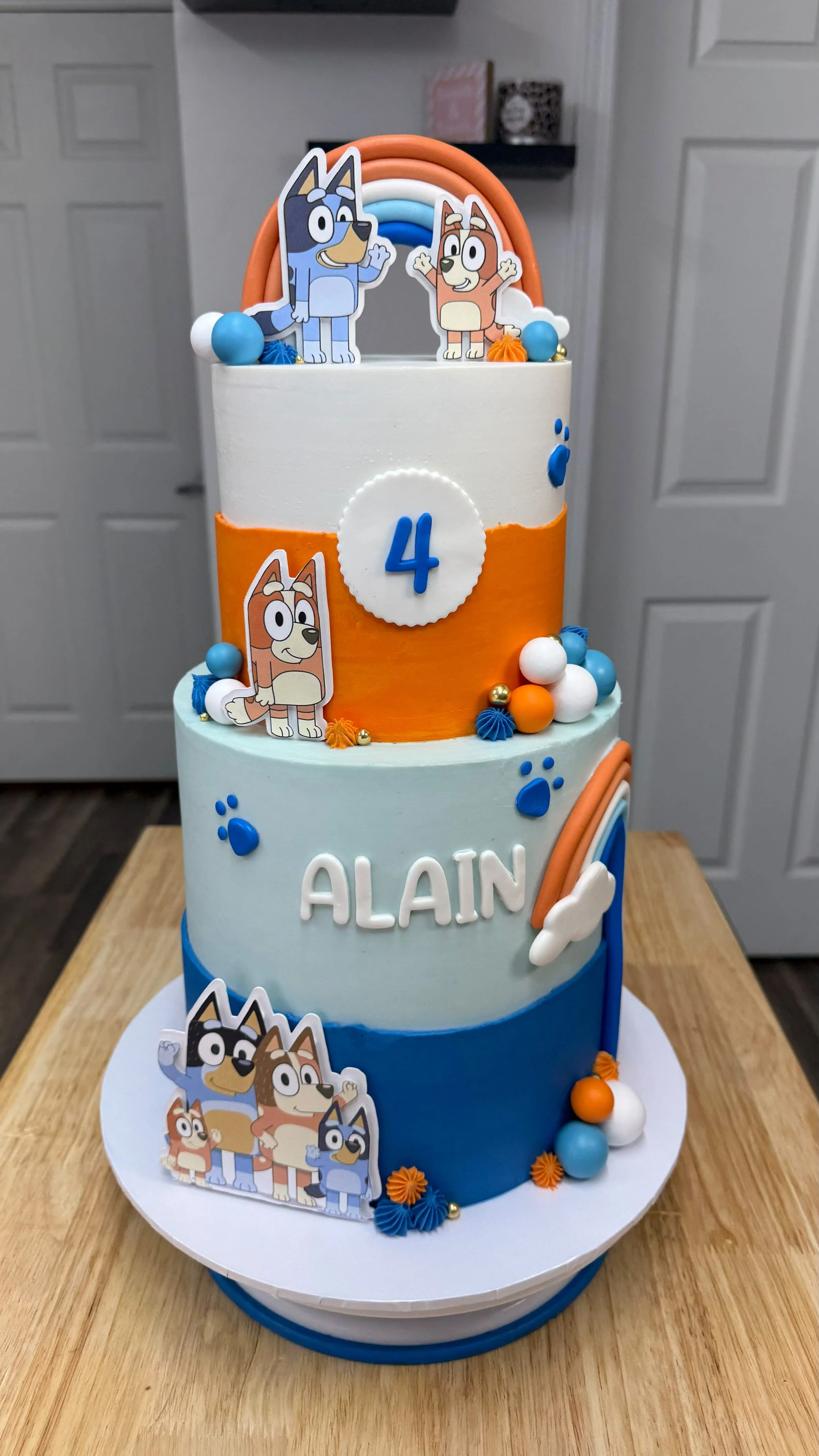 Pastel de cumpleaños de tres pisos decorado con personajes de dibujos animados, arcoíris y bolas de colores en tonos naranja, azul y blanco, con el nombre 'ALAIN' y el número 4.