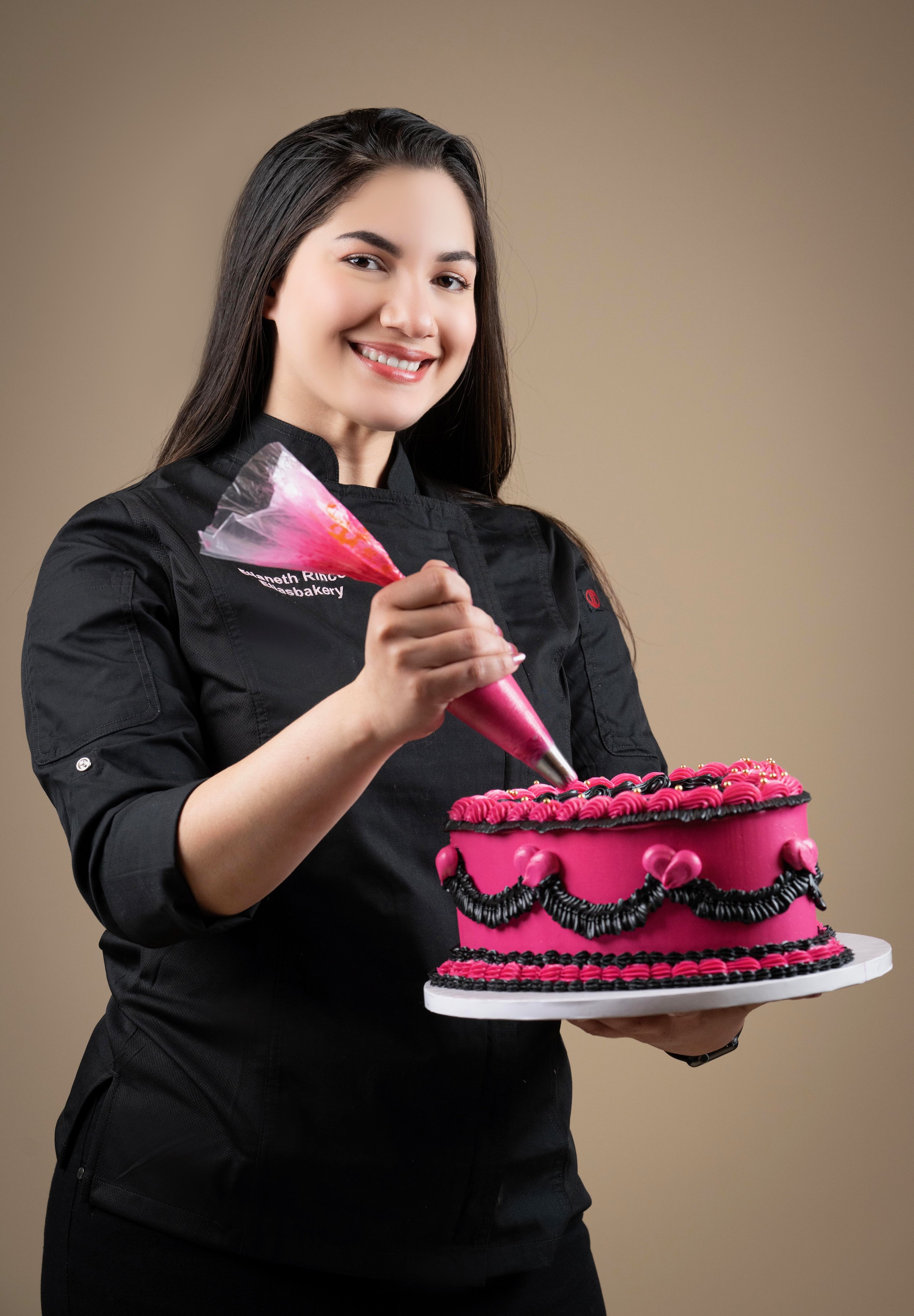 Mujer sonriendo, vestida con chaqueta negra de chef, sosteniendo un pastel decorado en tonos rosa y negro, usando una manga pastelera rosa para decorar el pastel.