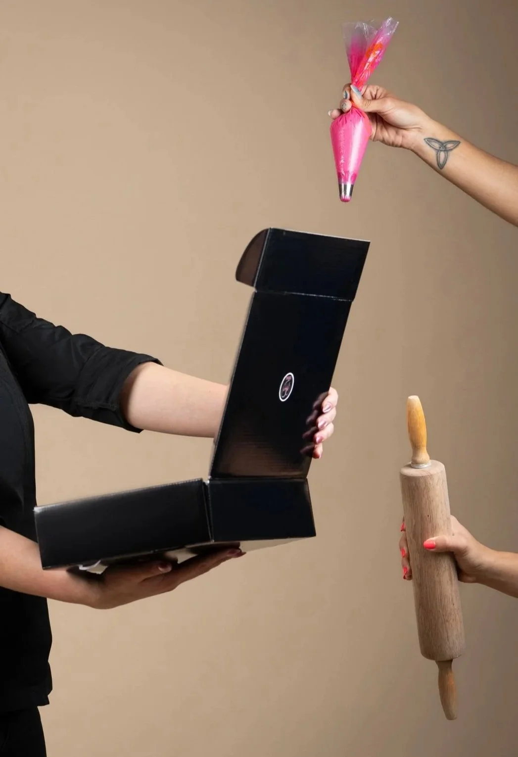 Manos sosteniendo una caja de pizza vacía, otra mano con un rodillo de madera, y una mano más con una bolsa de globo en forma de tubo con decoraciones rosadas, en frente de un fondo beige.