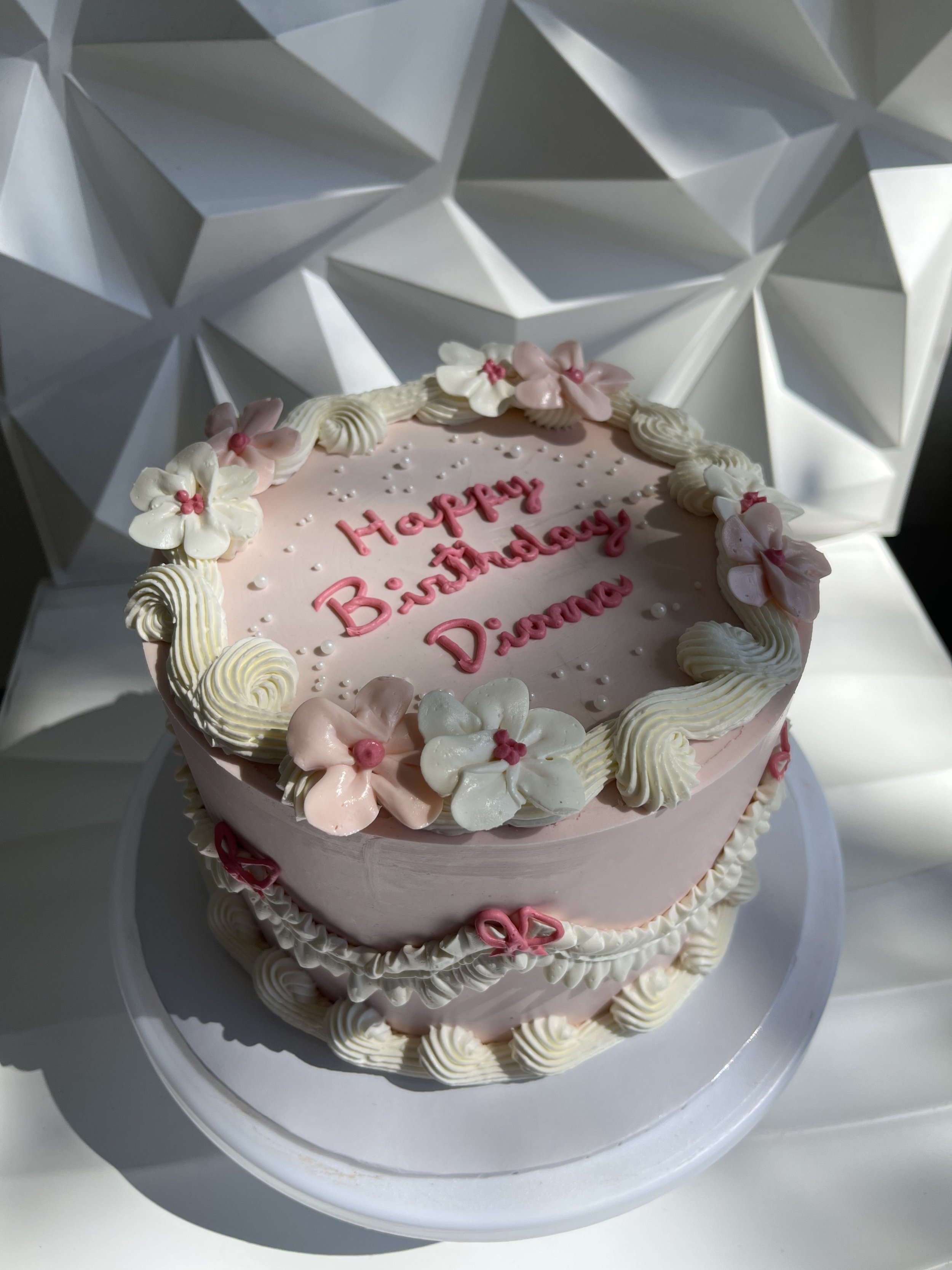 Pastel de colores pastel con decoraciones de flores y la frase 'Happy Birthday Diana' escrita en azúcar rosa, en un plato blanco.
