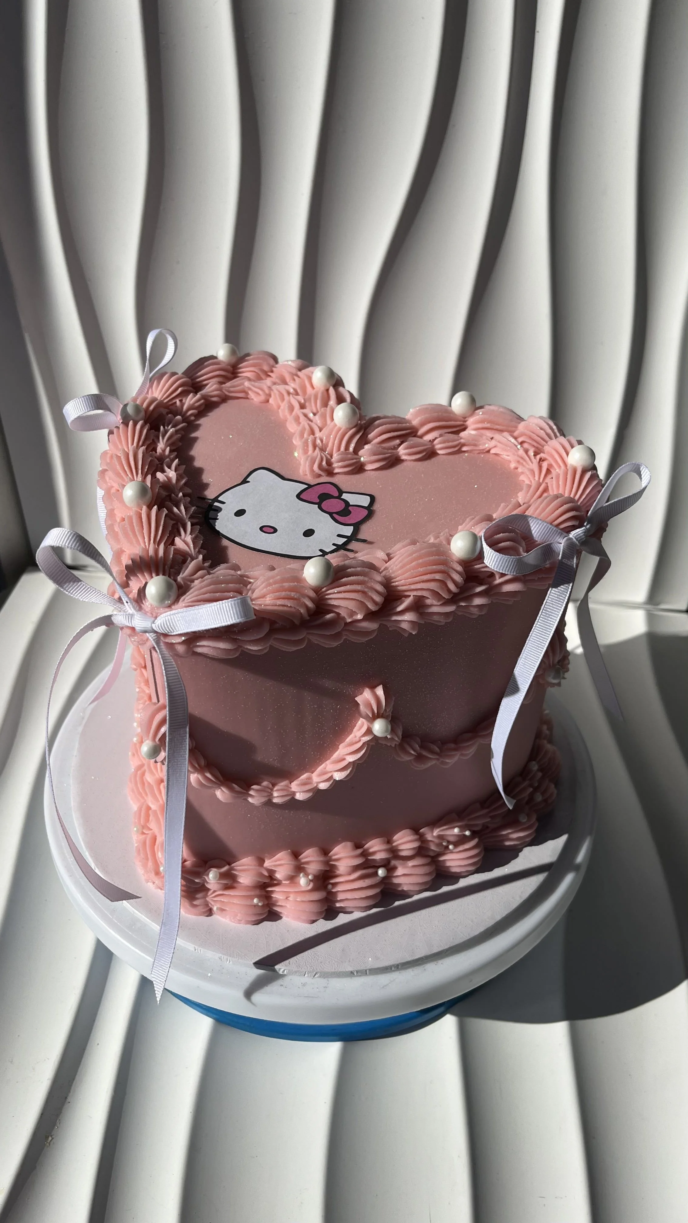 Pastel de cumpleaños en forma de corazón, decorado con rosa y blancas con la imagen de Hello Kitty en el centro, adornado con lazos de cinta y perlas decorativas.